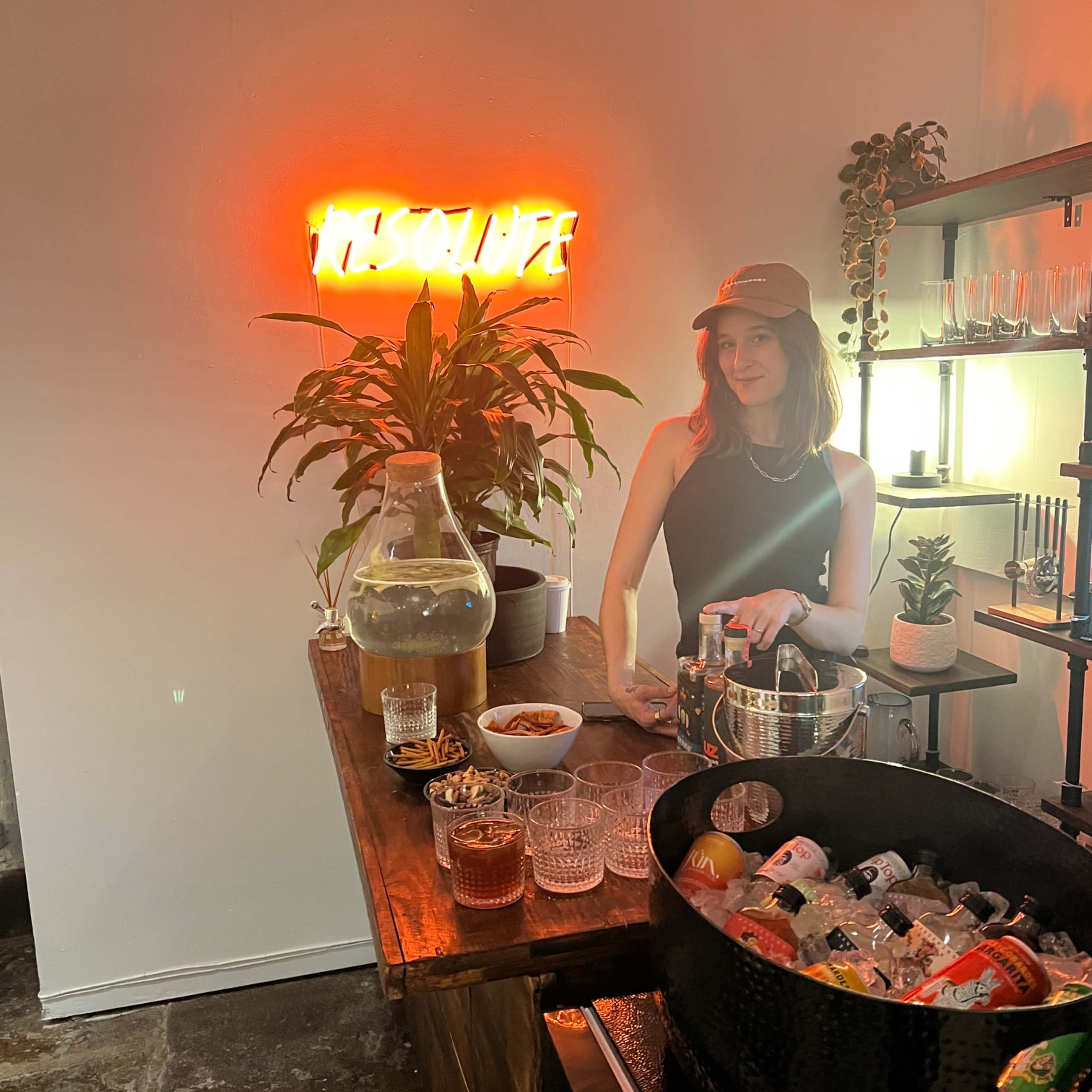 A woman stands at a bar counter with drinks and snacks, while a neon sign reads "ABSOLUTE" on the wall behind her.