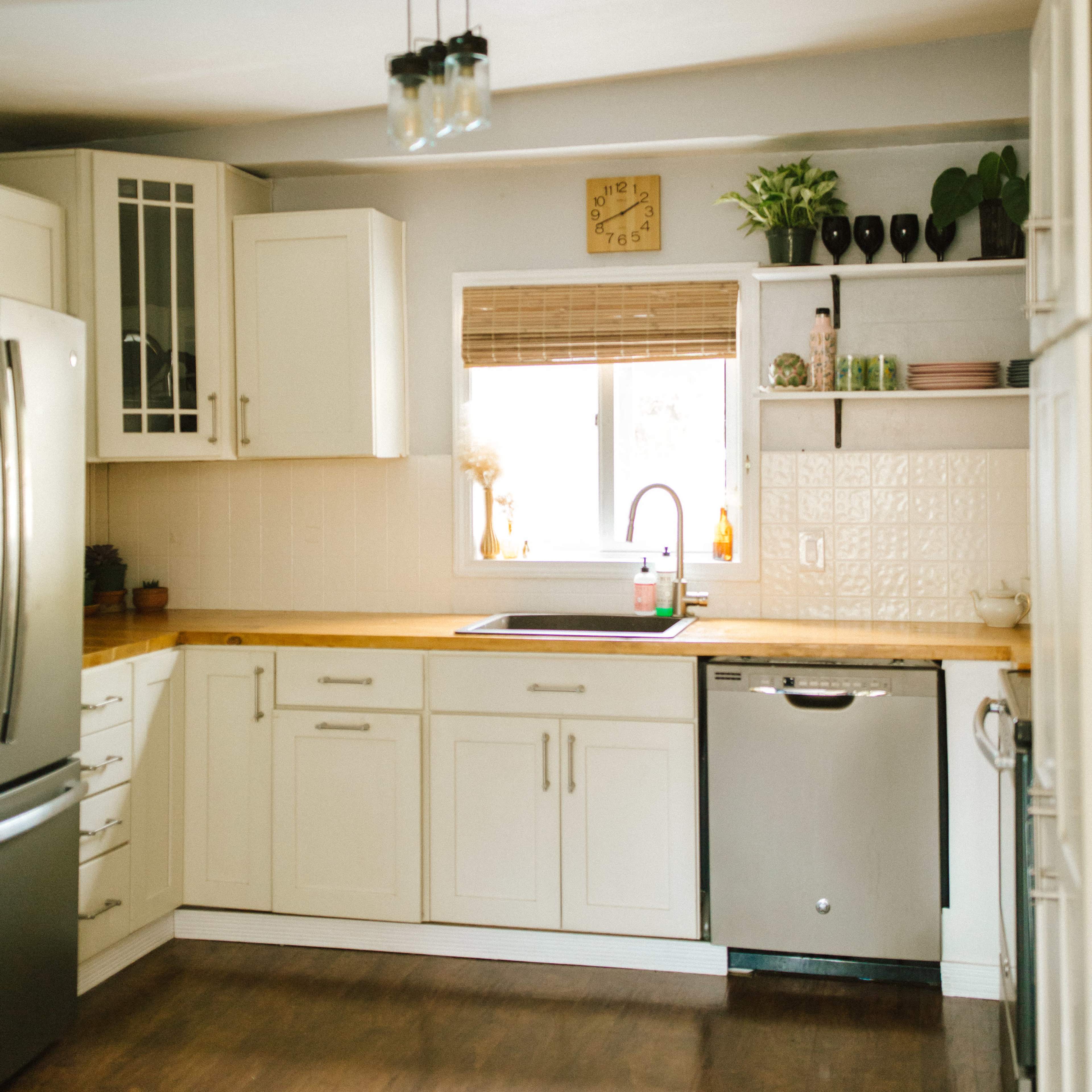 A bright kitchen features cream-colored cabinets, a stainless steel refrigerator, and a sink below a window with a bamboo shade.
