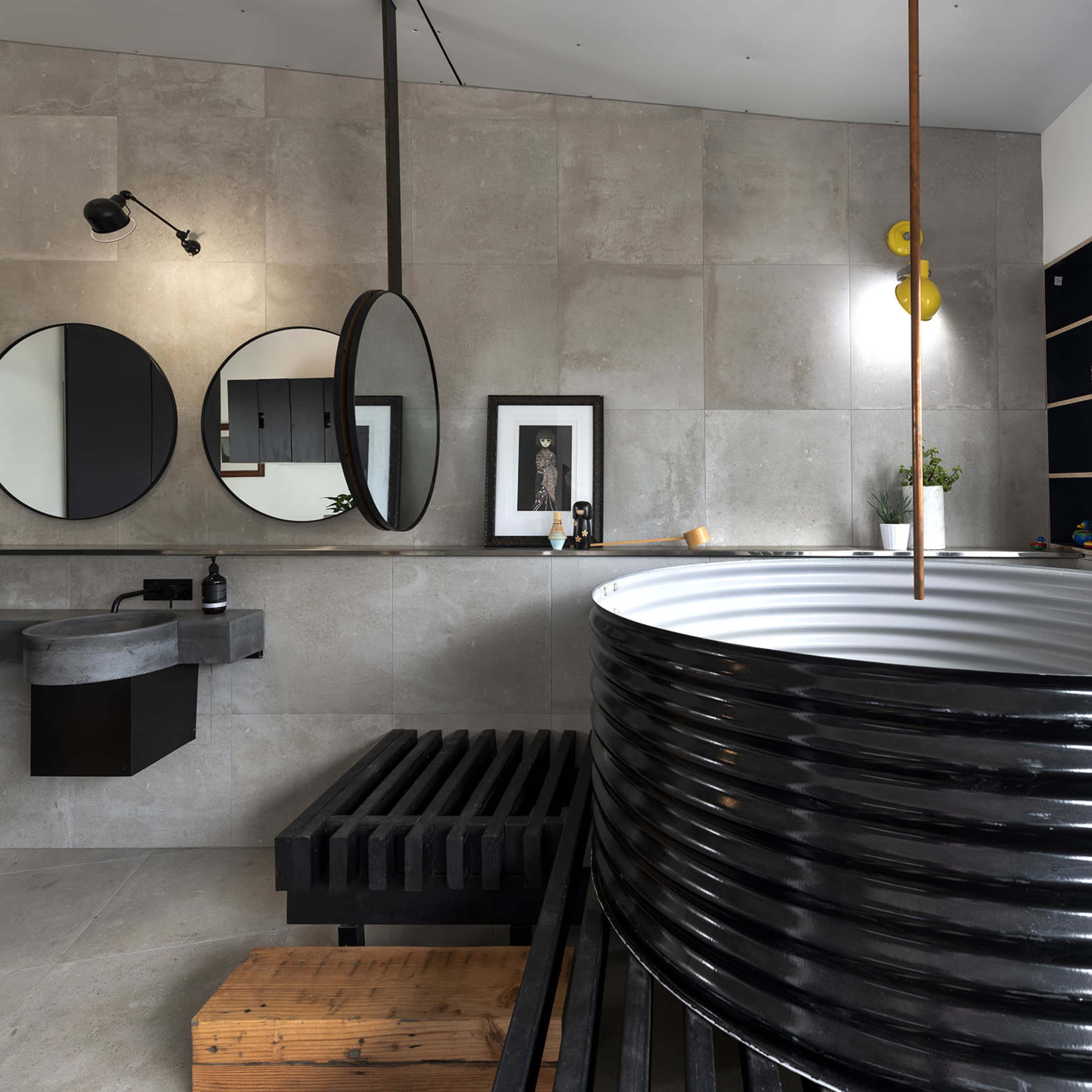 A modern bathroom featuring a round black metal bathtub, a wall-mounted sink, two circular mirrors, and minimalist decor.