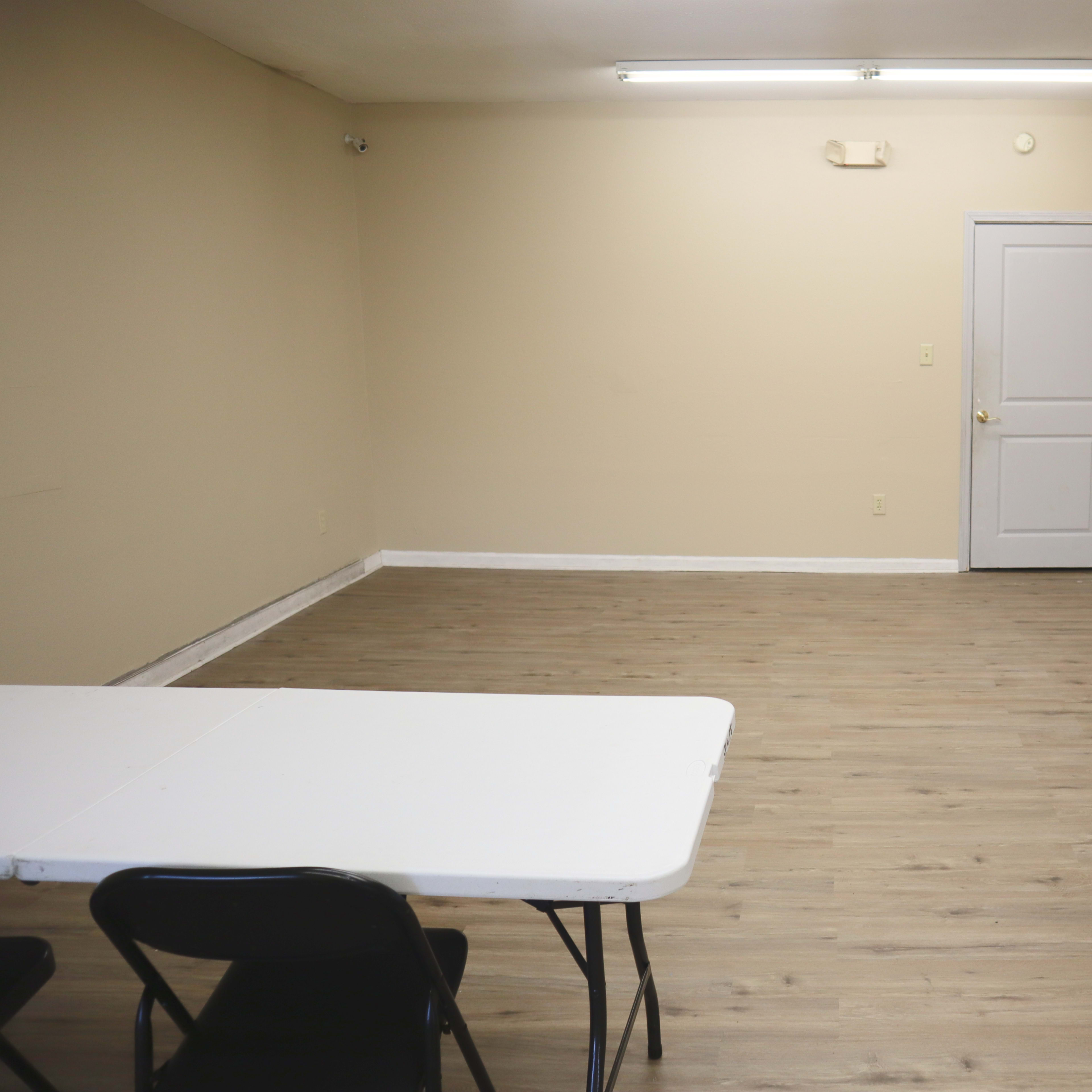 A spacious, empty room with a light brown wooden floor, a white folding table, and a closed door.