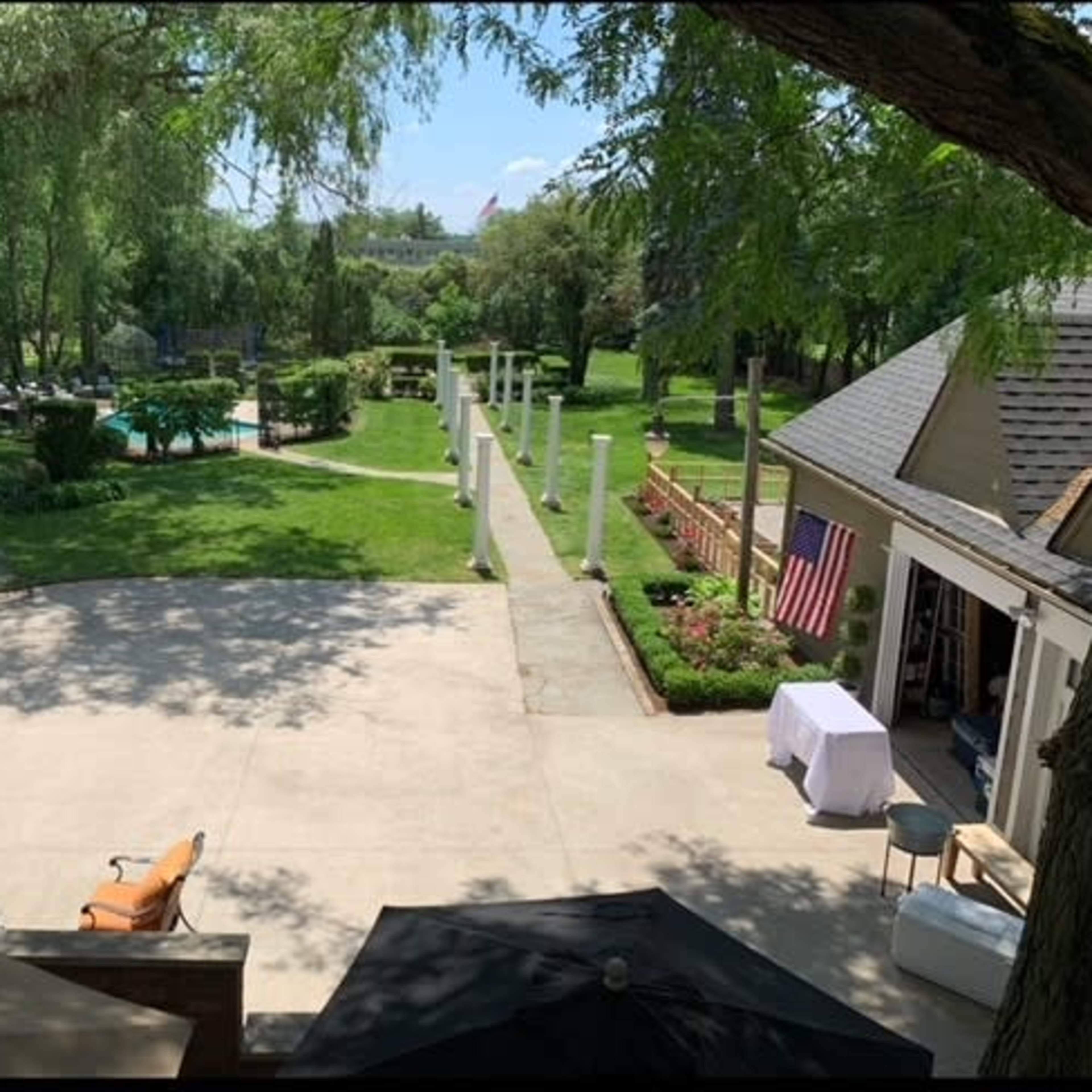 A spacious backyard with a concrete patio, a swimming pool, landscaped gardens, and several standing columns.