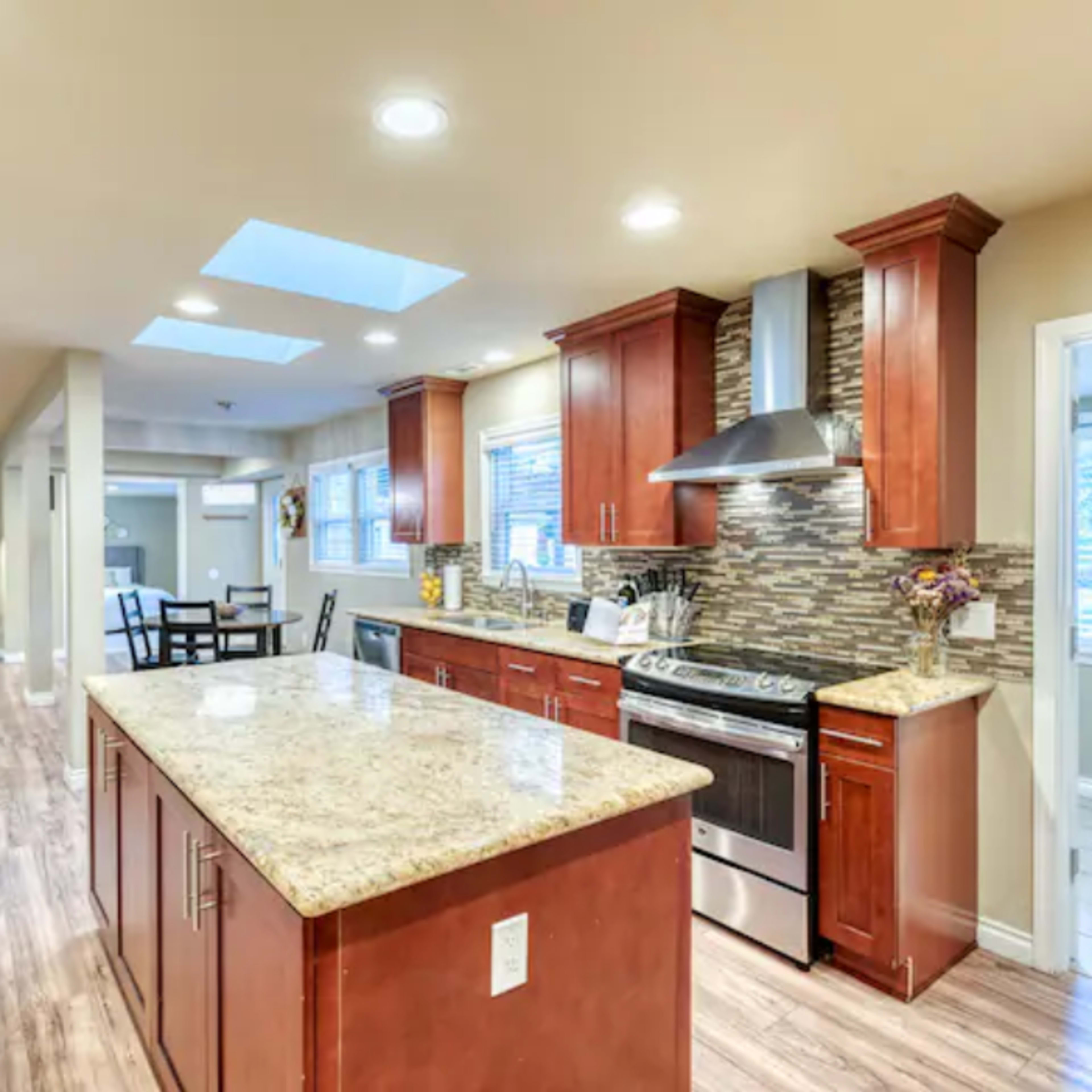 The kitchen features wooden cabinets, a granite countertop island, stainless steel appliances, and natural light from skylights above.