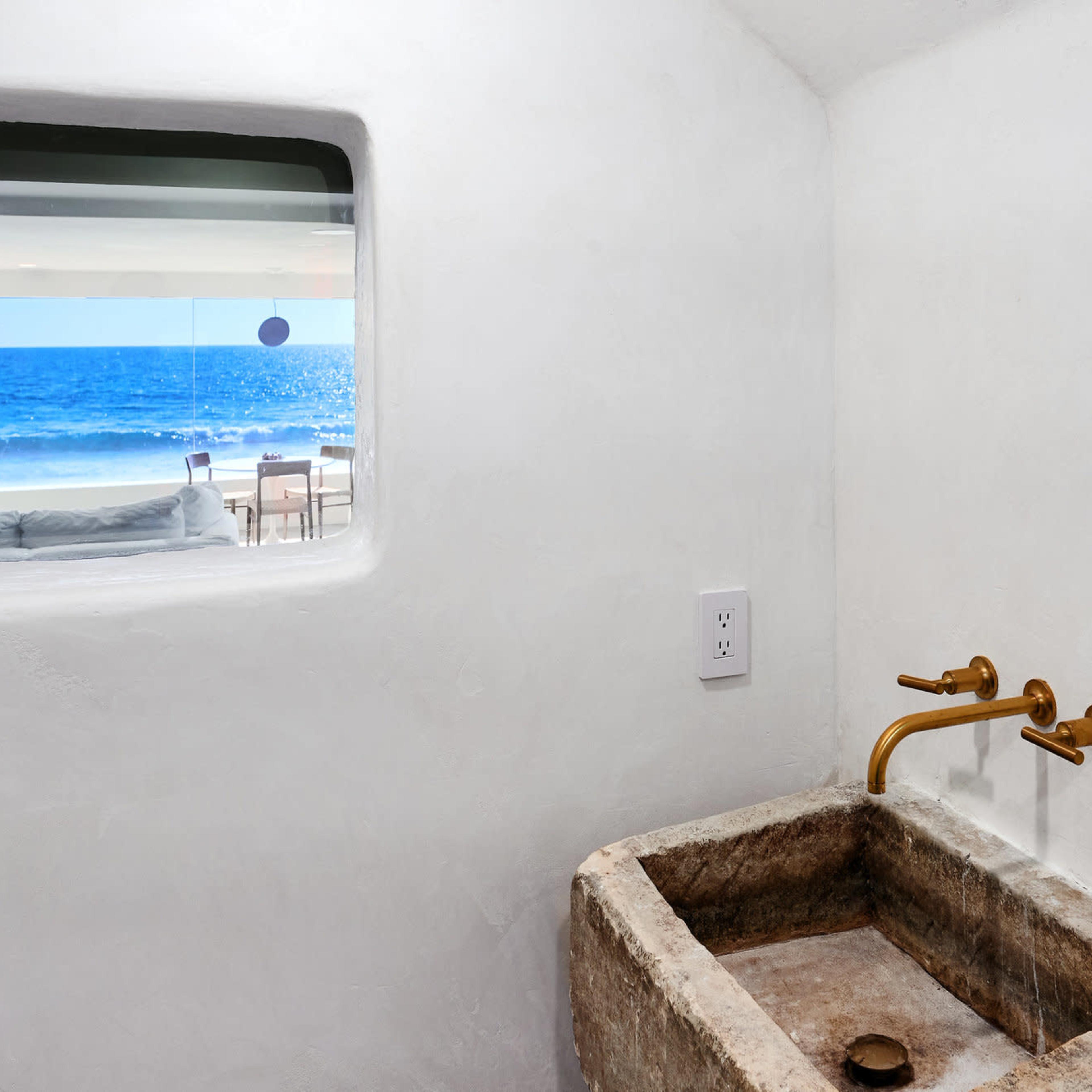 A stone sink is mounted against a white wall, with a window showcasing a view of the ocean and outdoor seating area.