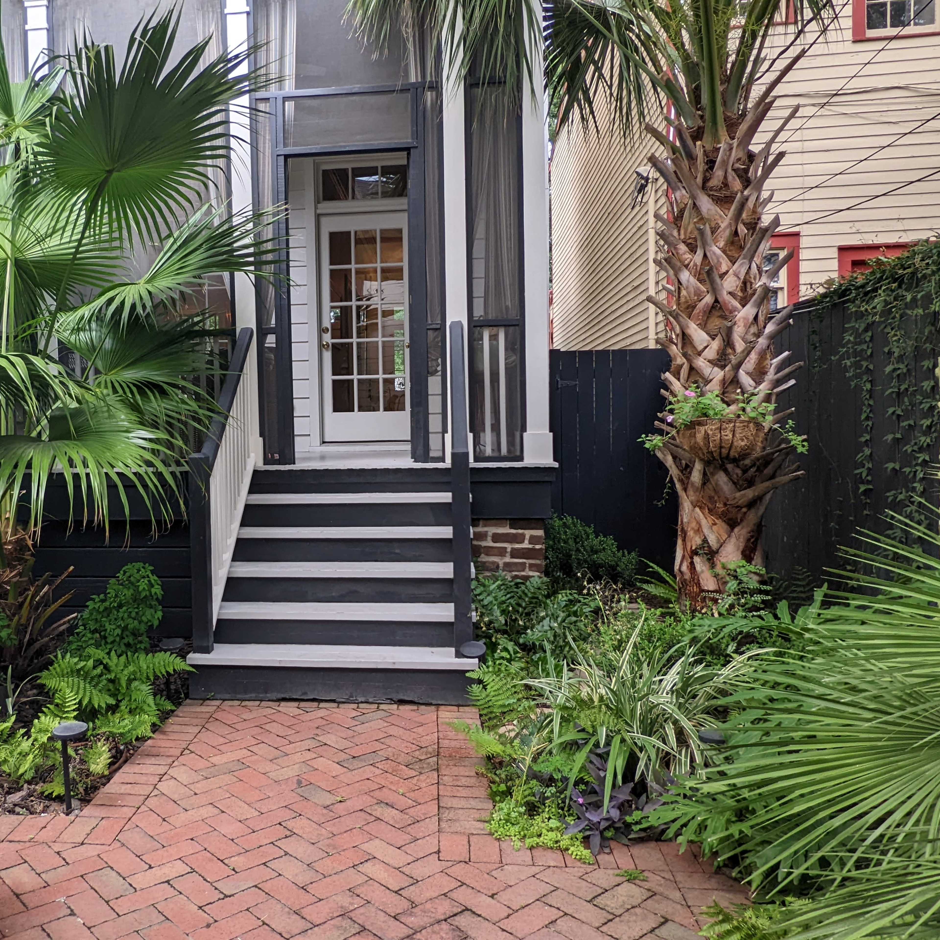 A set of steps leads to a glass door surrounded by lush greenery and palm trees in a brick-paved courtyard.