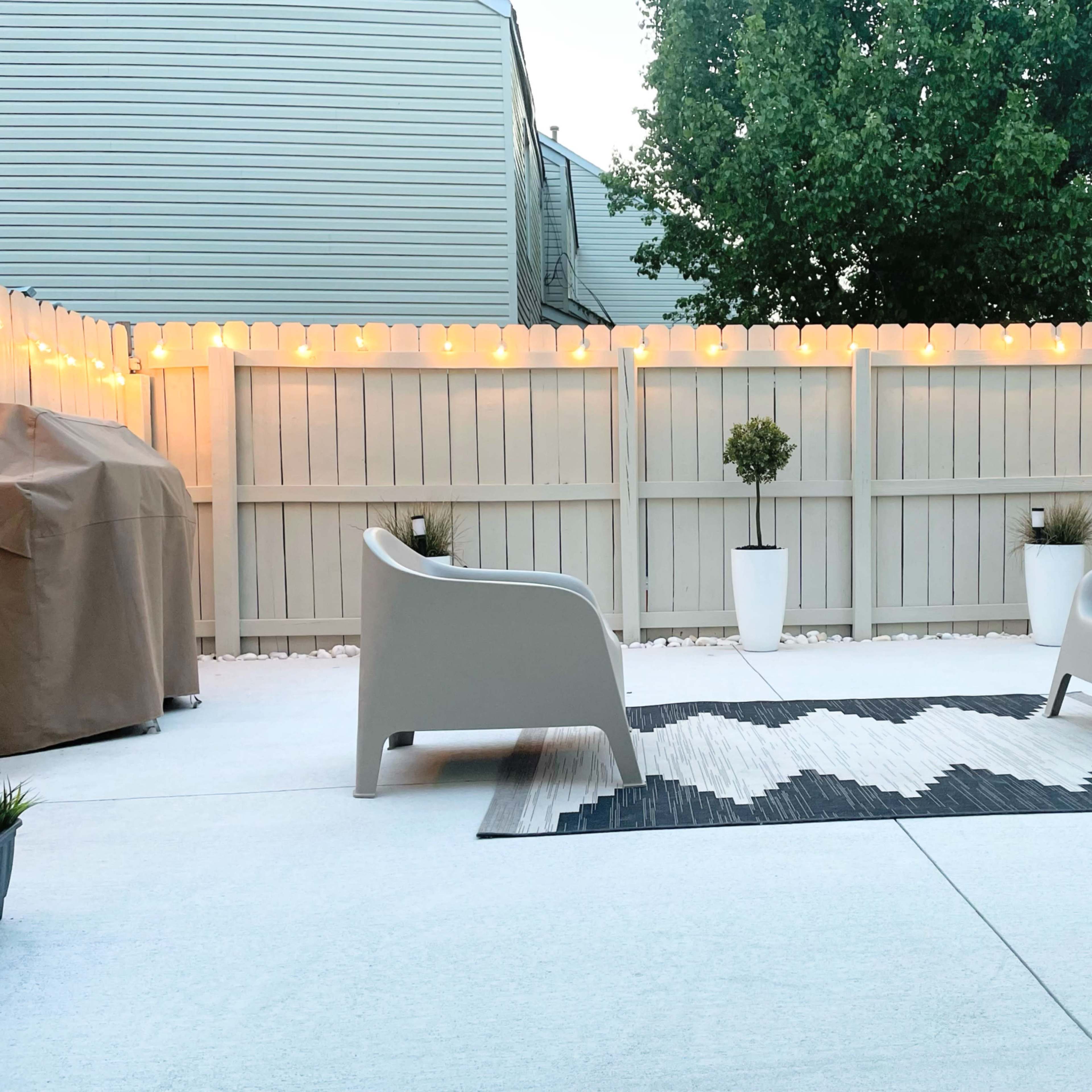 The patio features two gray chairs positioned on a rug, with potted plants and string lights illuminating the enclosed space.