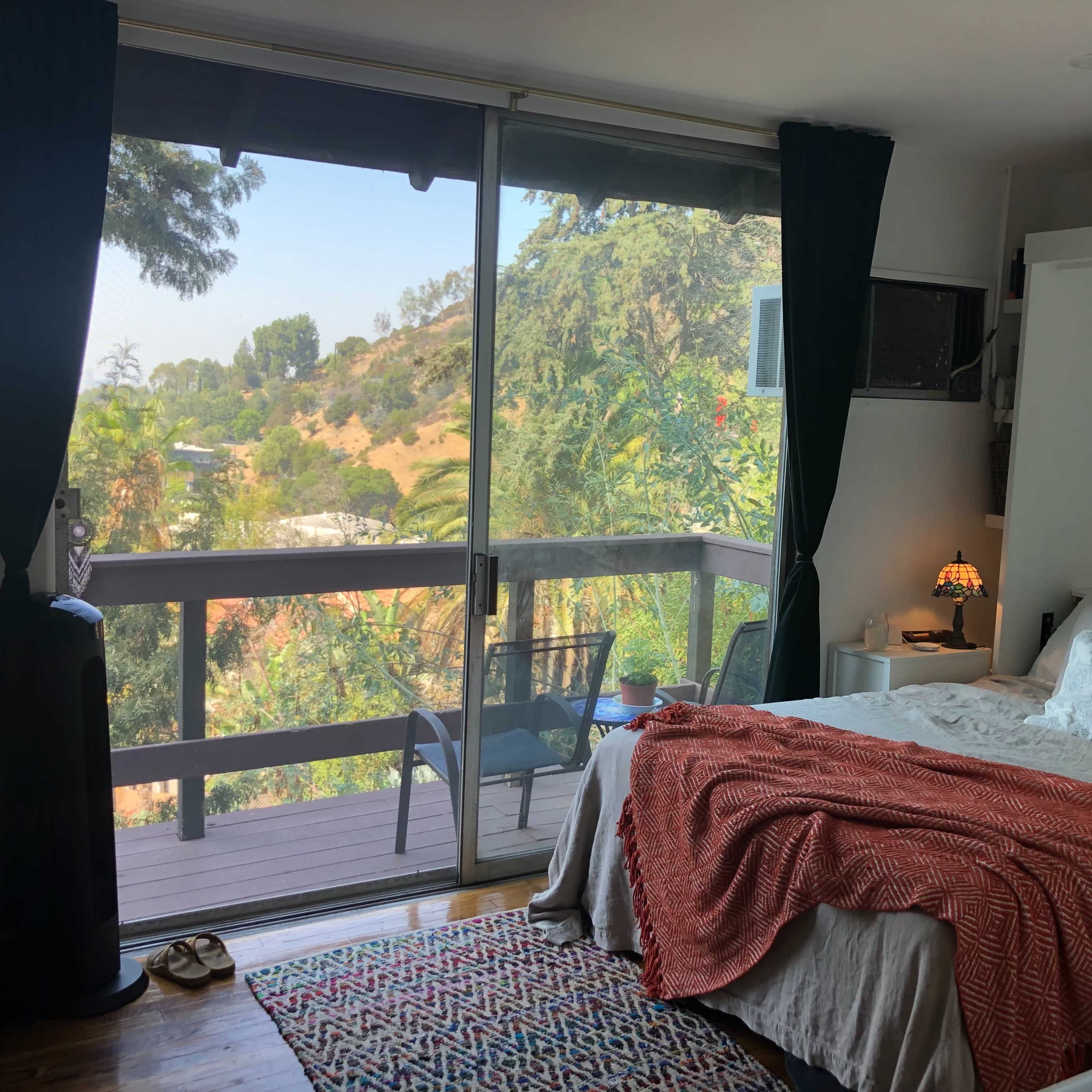 A bedroom features a bed with a patterned blanket, a TV mounted on the wall, and large sliding glass doors that open to a view of green hills.