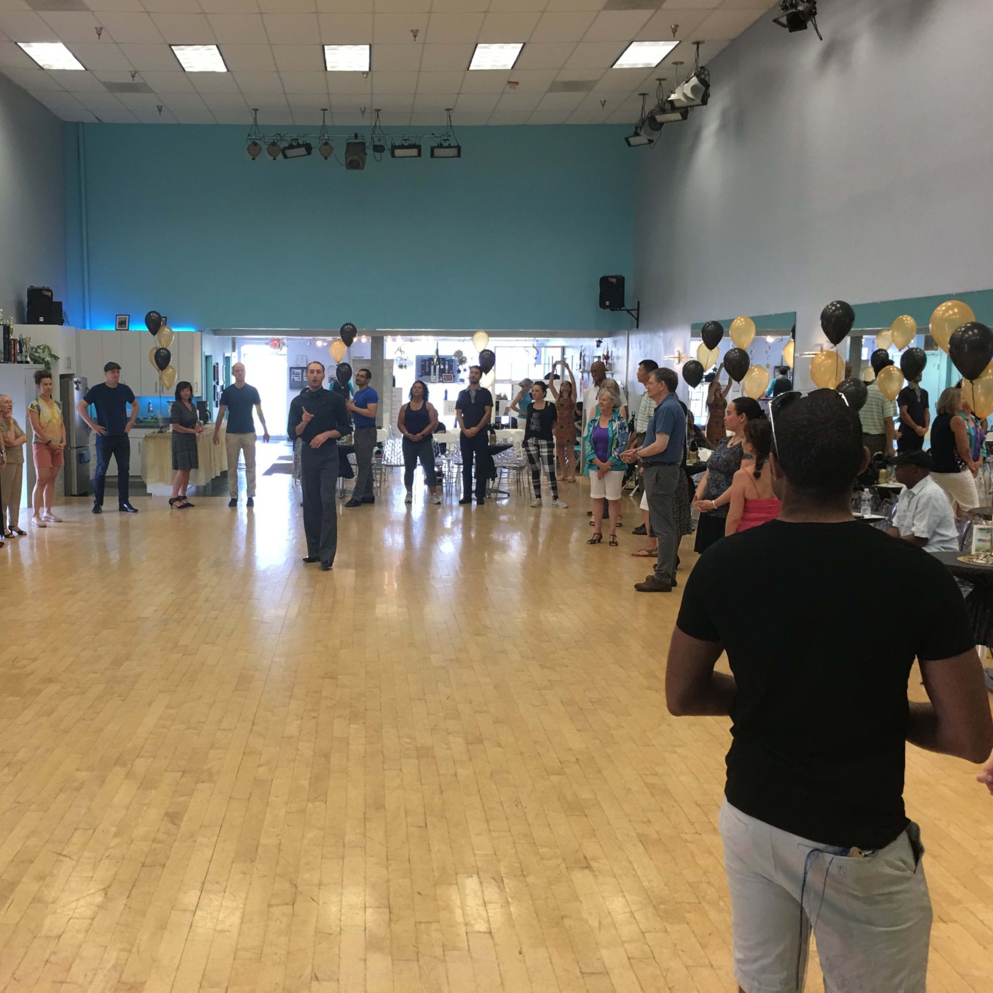 A large group of people stands in a circle inside a spacious dance studio, while a speaker addresses them from the center.