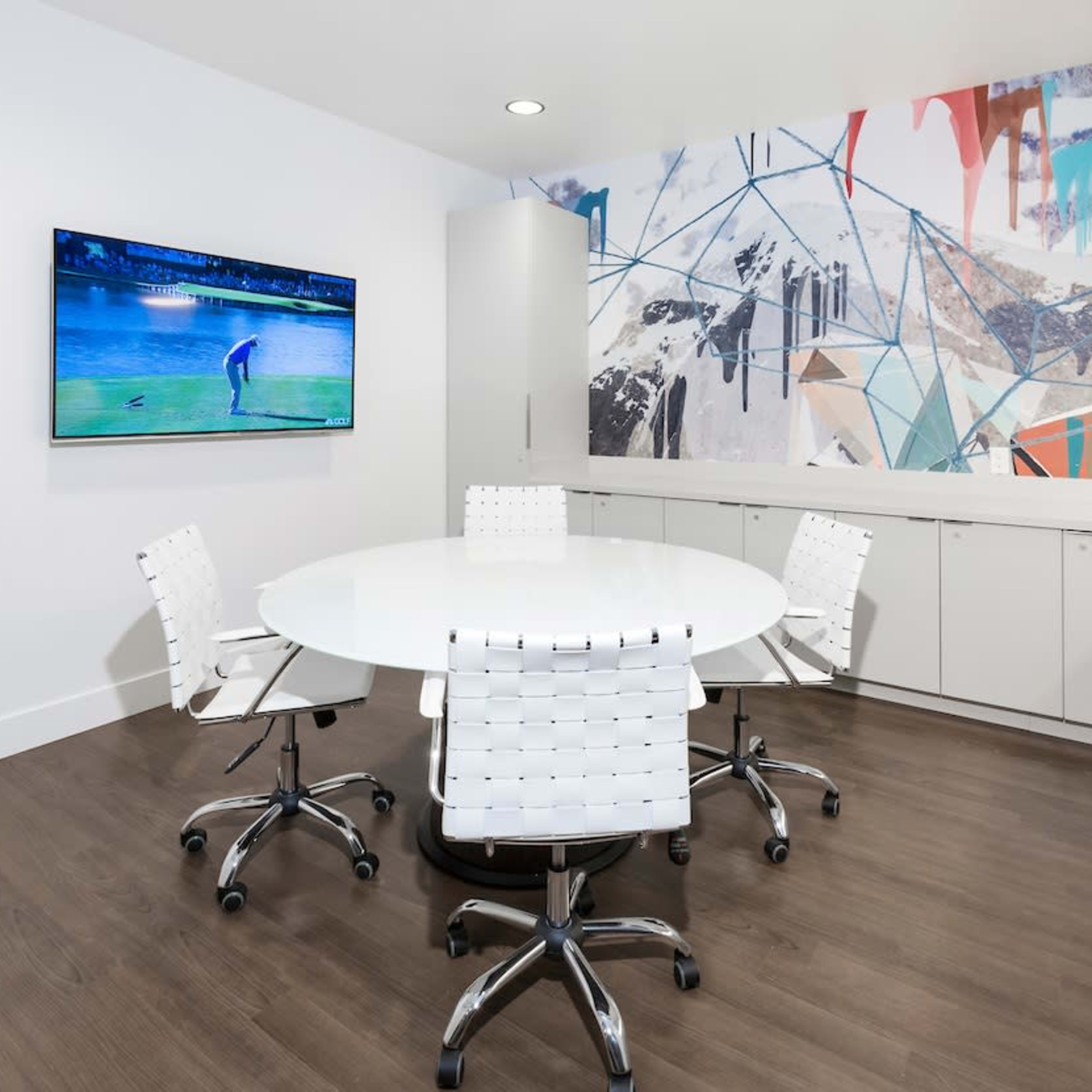 A modern conference room with a circular table, four white chairs, a television on the wall displaying a golf scene, and a colorful abstract mural.