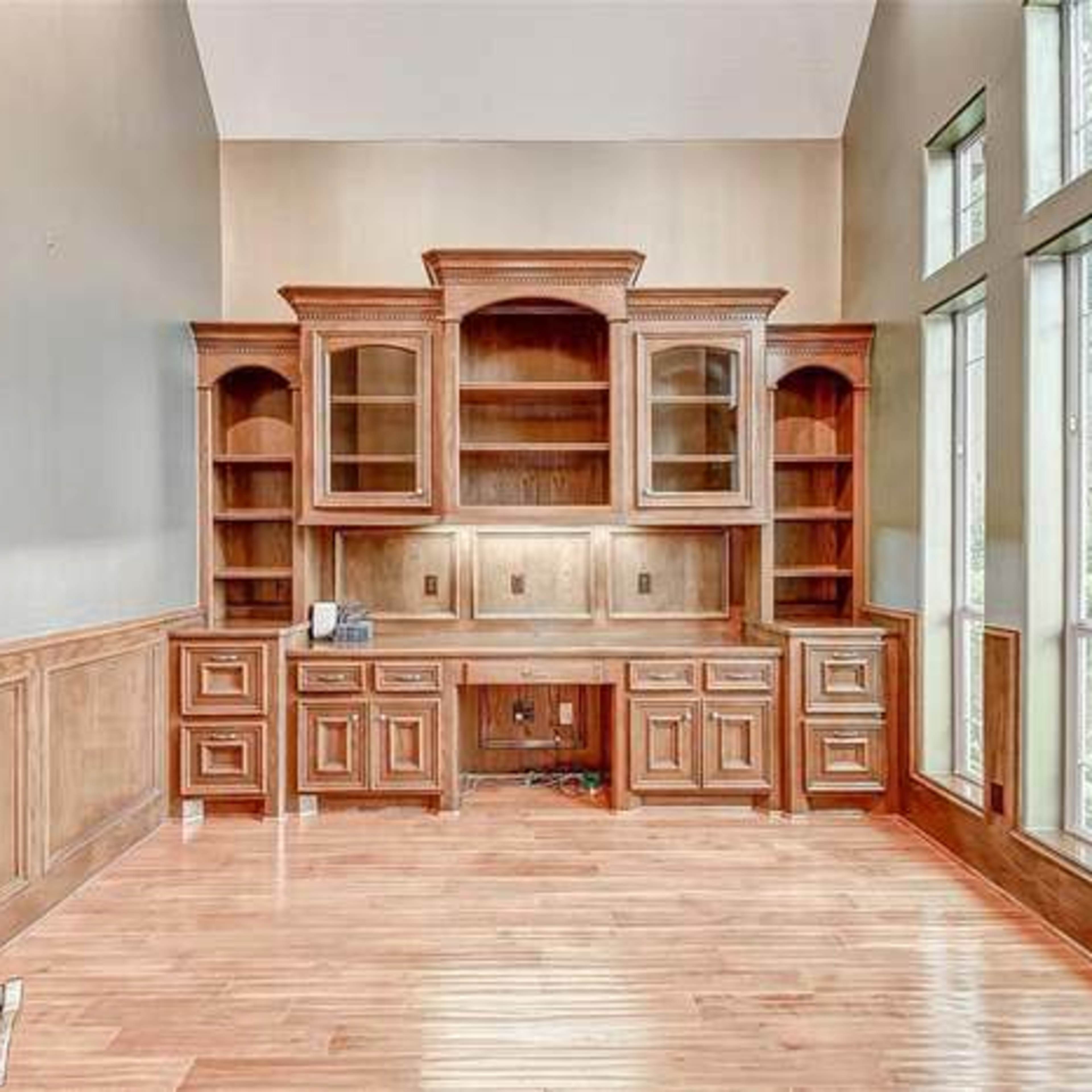 A spacious room with a wooden built-in desk and shelves, large windows providing natural light, and polished wooden flooring.