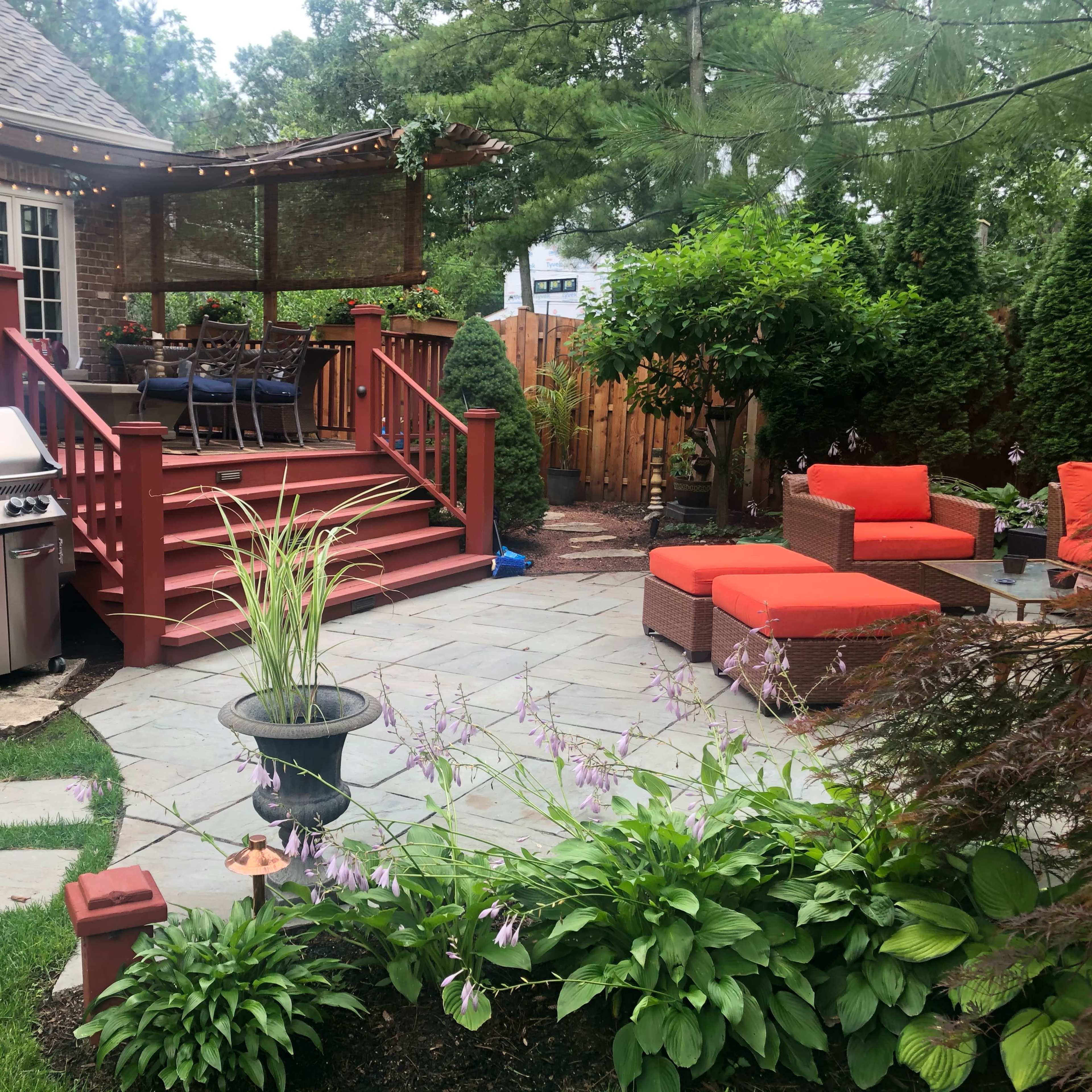 A landscaped backyard featuring a wooden deck with seating, a gas grill, and an outdoor stone patio surrounded by greenery and flower beds.