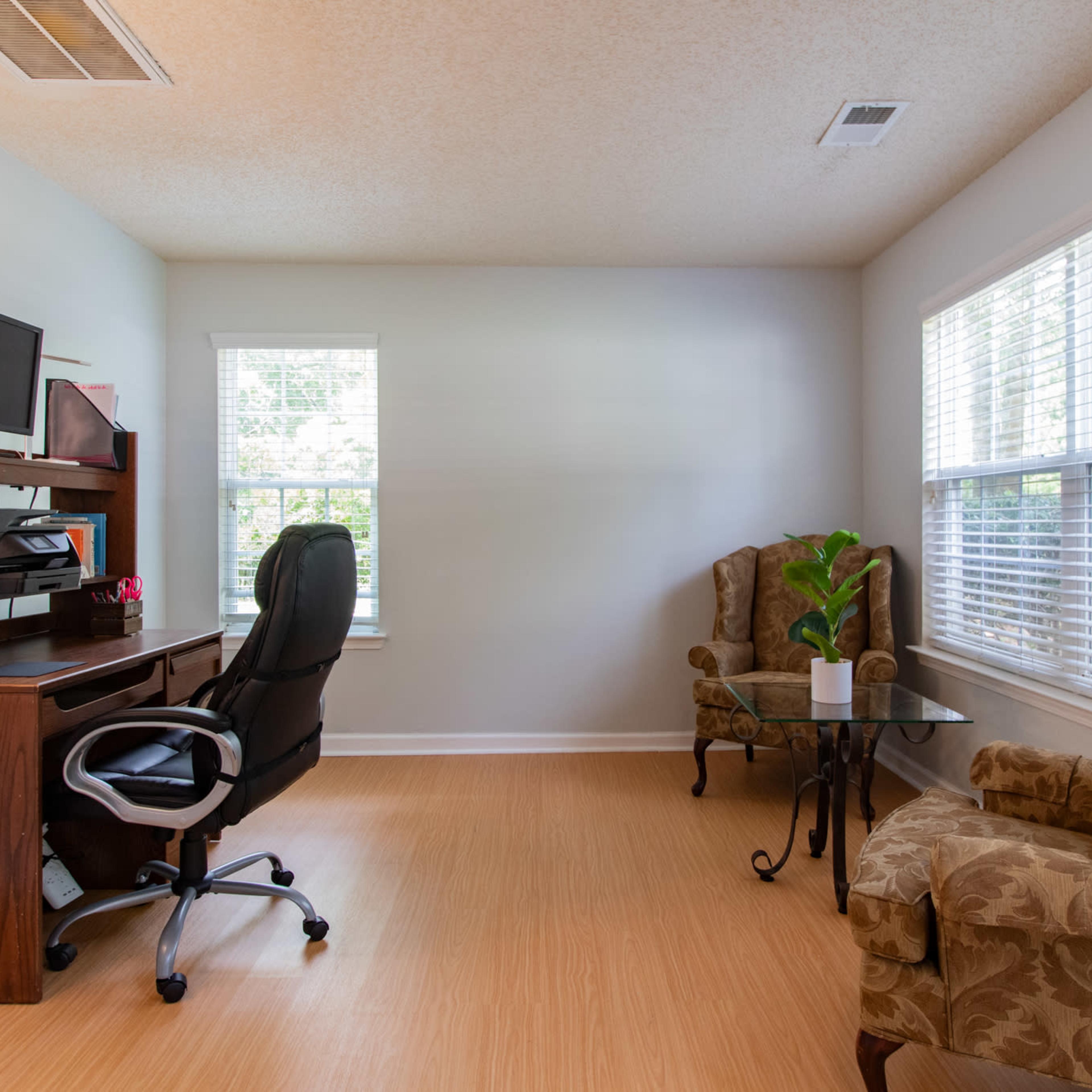 A home office with a wooden desk and chair, a computer, two upholstered armchairs, and a small table by a window.