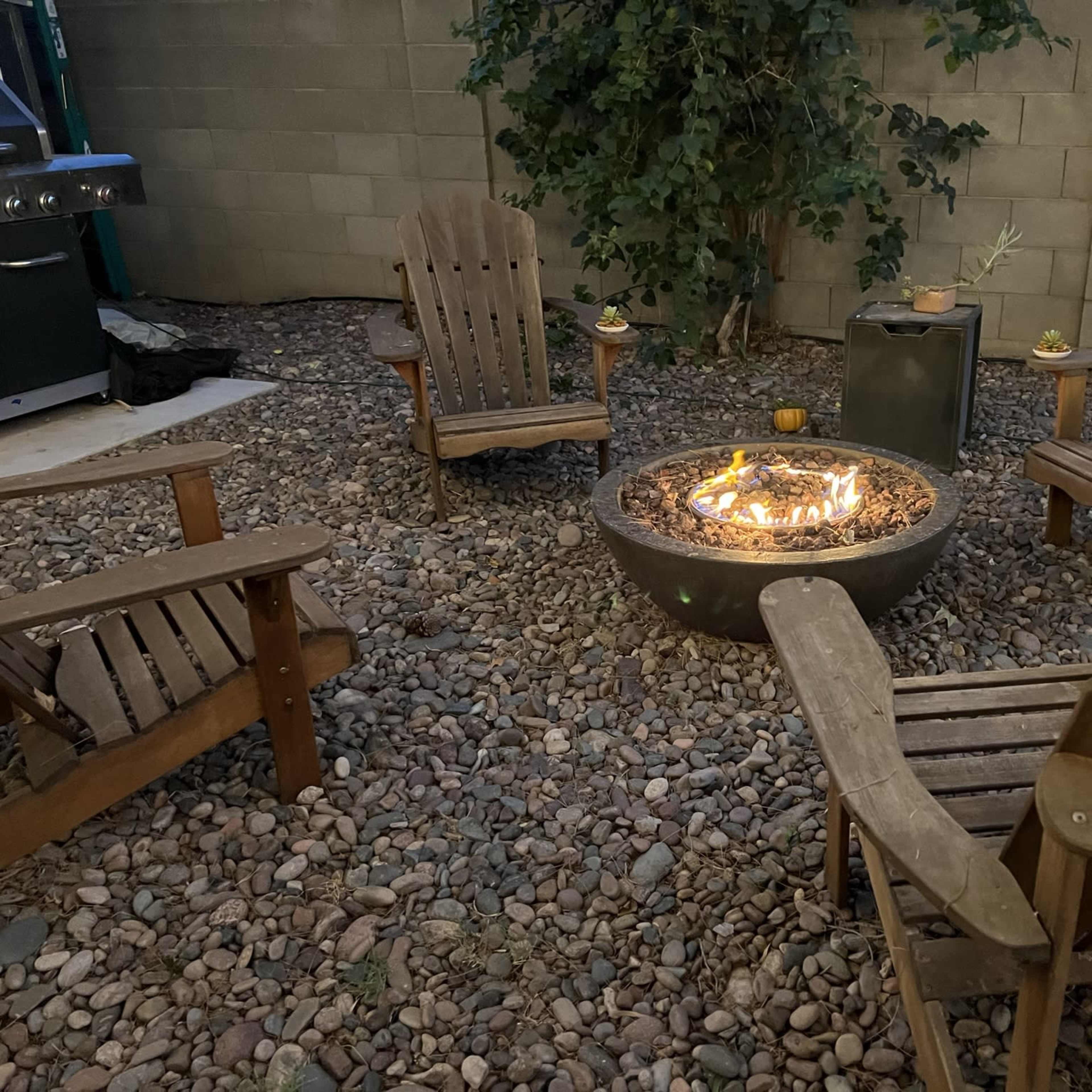 A fire pit is surrounded by four wooden chairs on a rocky surface in a backyard setting.