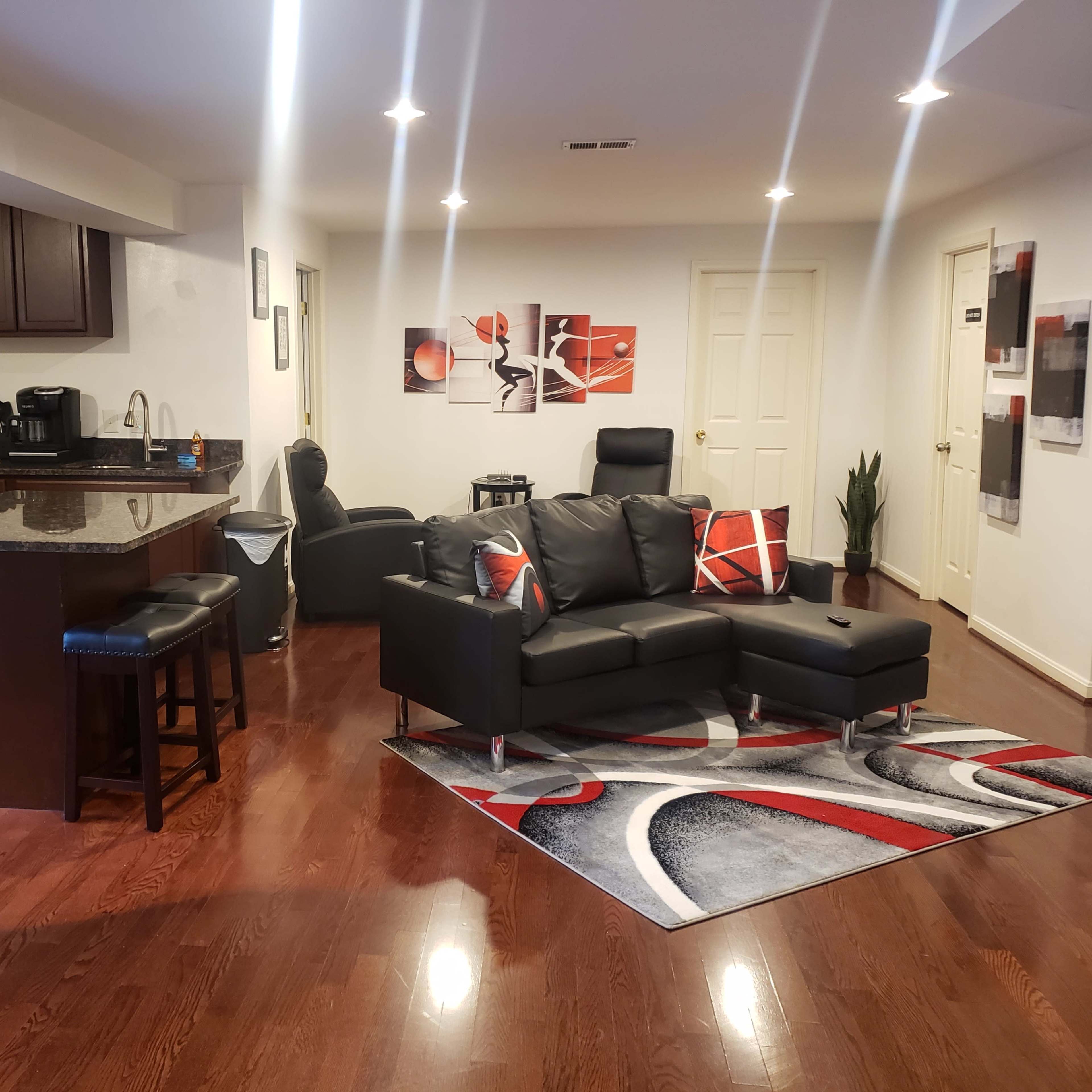 A modern open-concept living space featuring a kitchen with dark wood cabinets, stainless steel appliances, and a living area with a black sectional sofa and decorative rug.
