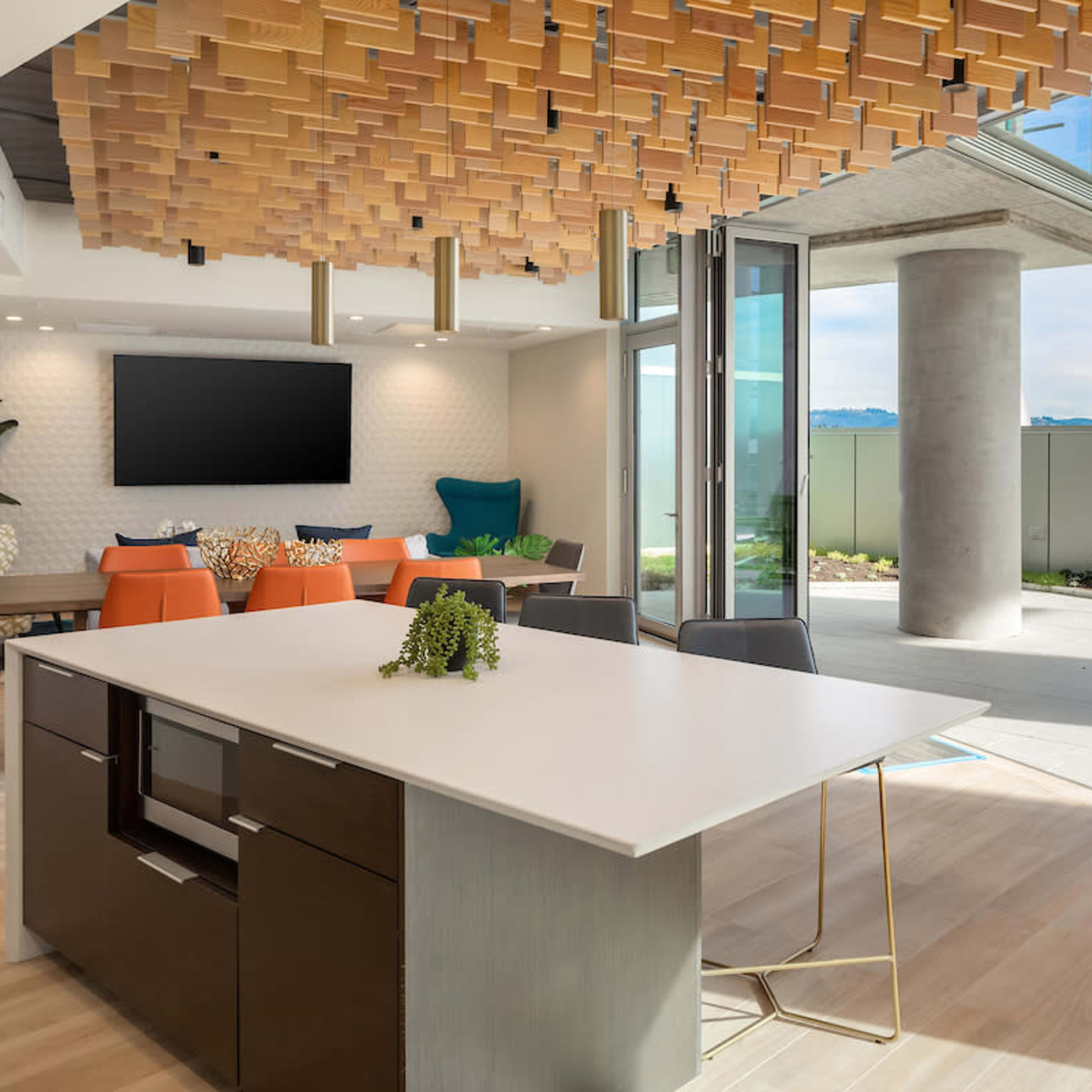 A modern open-concept lounge area with a large island table, stylish seating, and sliding glass doors leading to a patio with greenery.