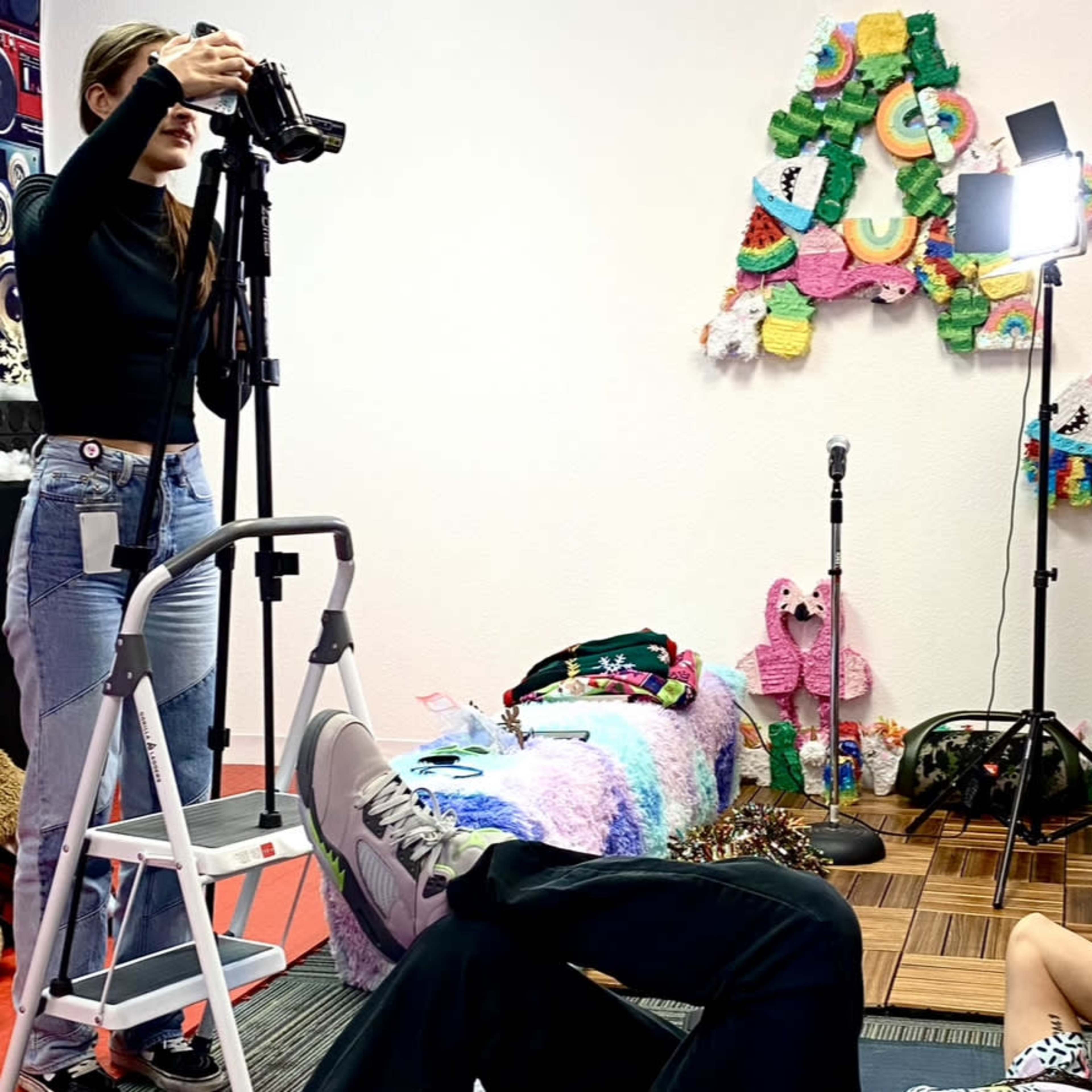 A photographer stands on a ladder, taking pictures of a person lying on the floor in a decorated room with colorful wall art and a soft light.