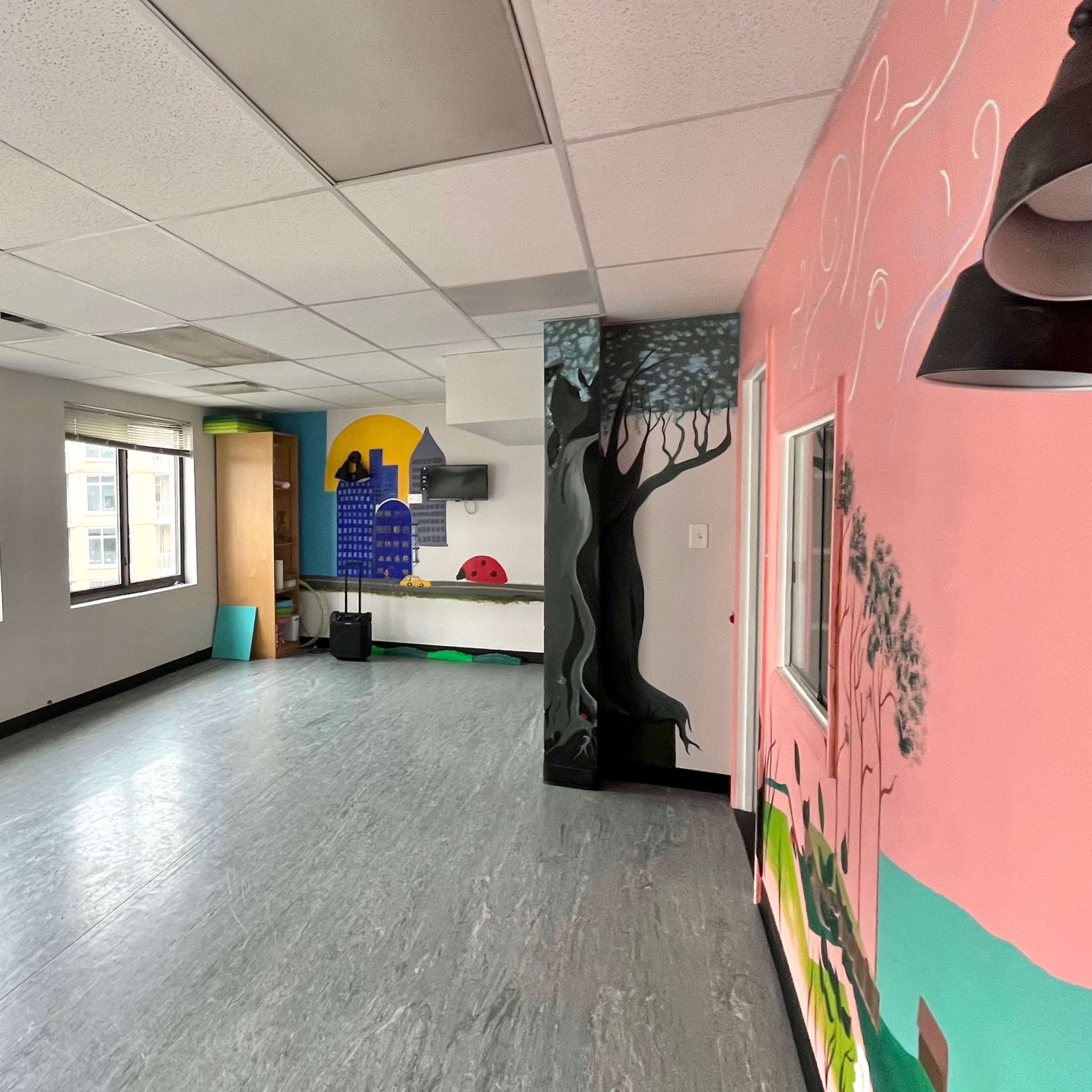 A brightly colored room with large windows, featuring a mural on one wall and a partially visible kitchenette area.