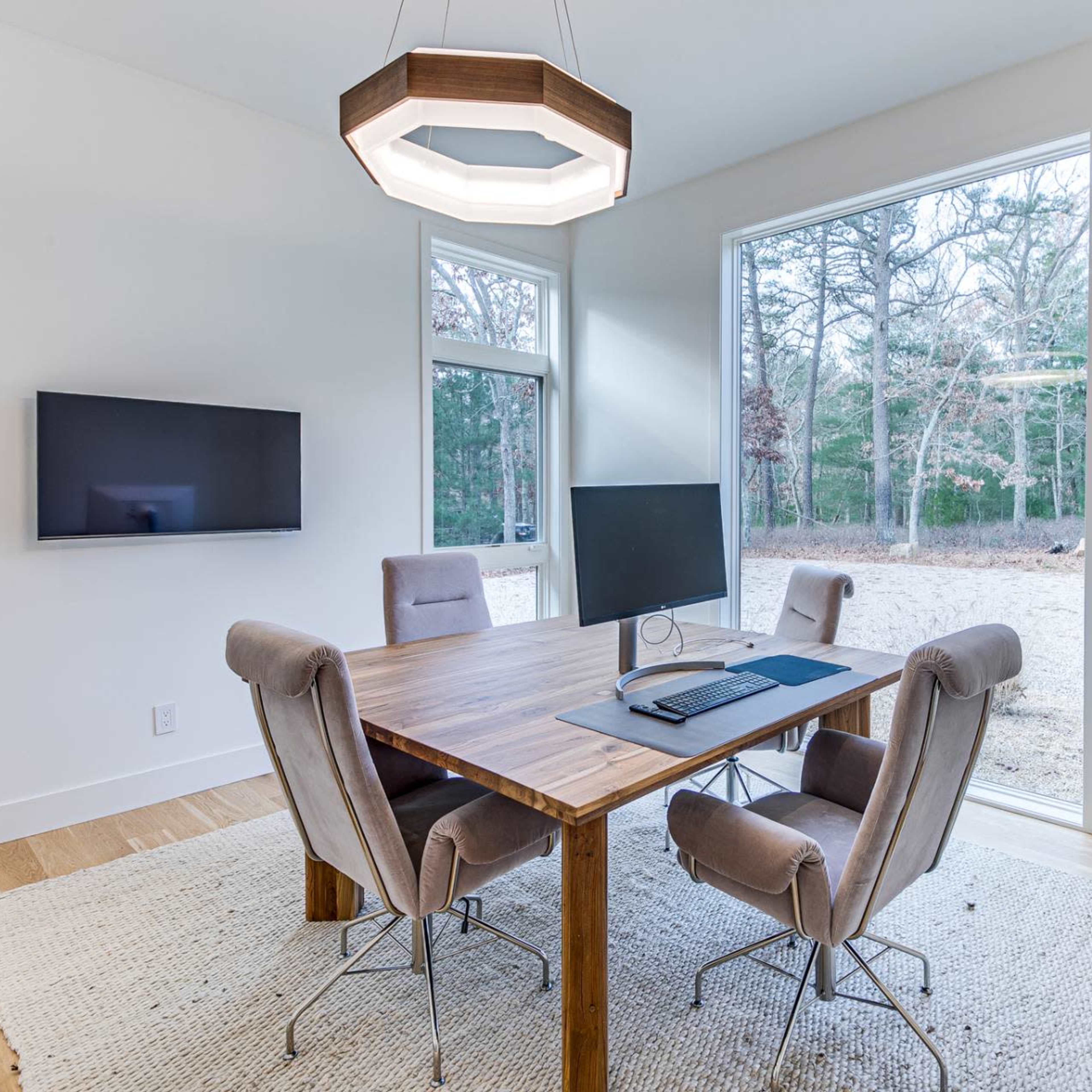 A wooden table with a computer and chairs is set in a bright, minimalist room with large windows overlooking a wooded area.
