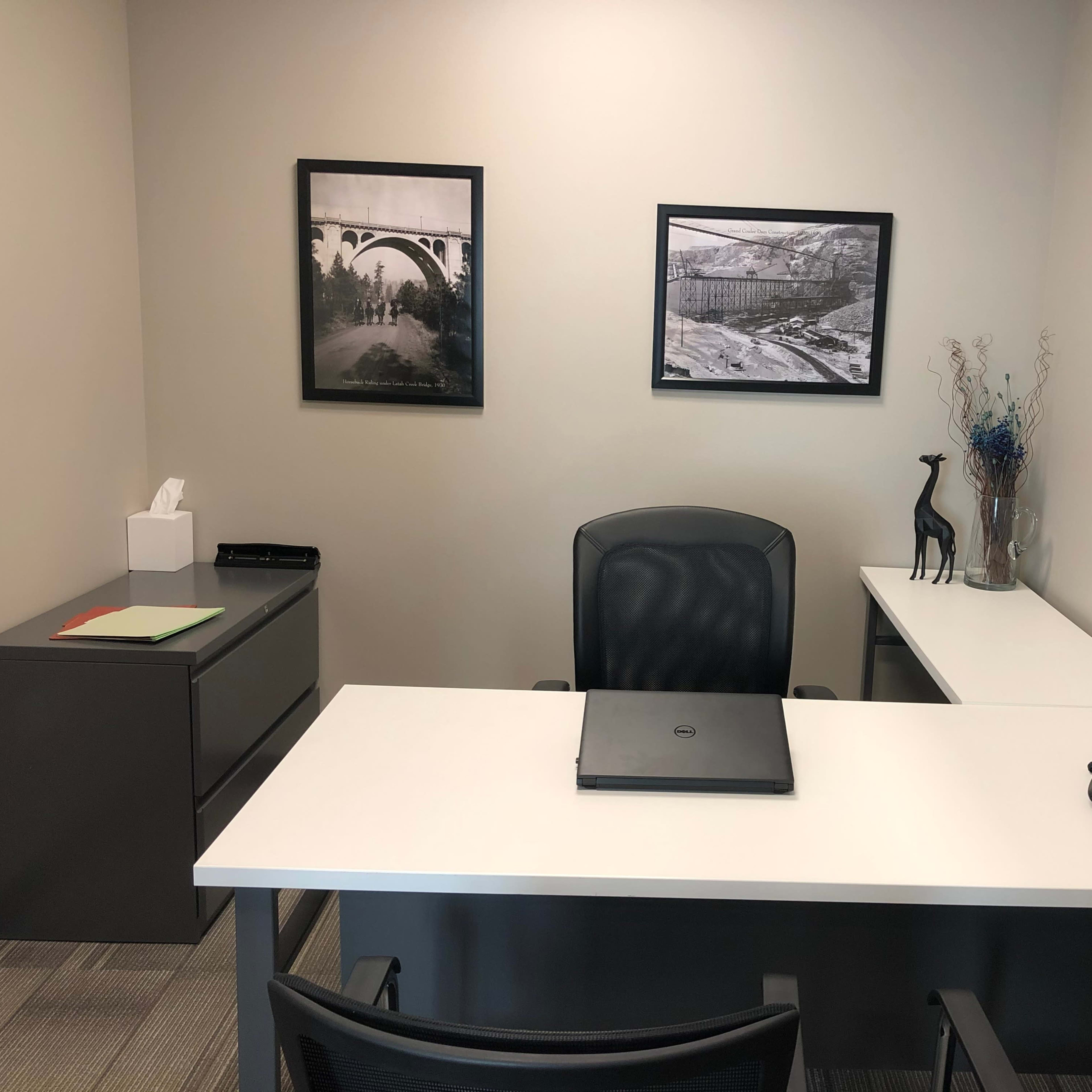 A modern office space features a desk with a laptop, a telephone, two framed black-and-white photographs on the walls, and a small decorative giraffe on the tabletop.