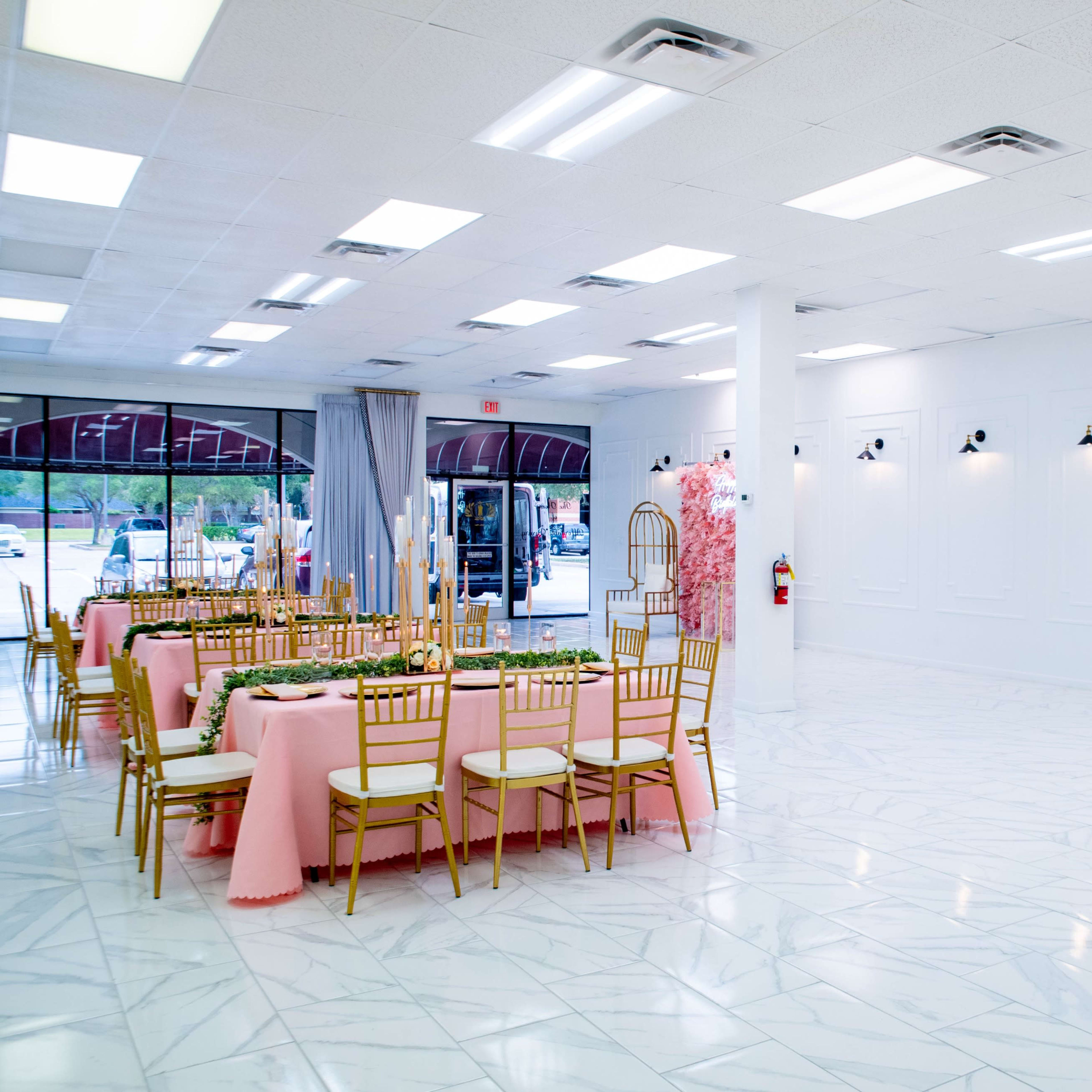 A banquet hall with a pink tablecloth-covered table set for an event, surrounded by gold chairs, and featuring white walls and marble flooring.