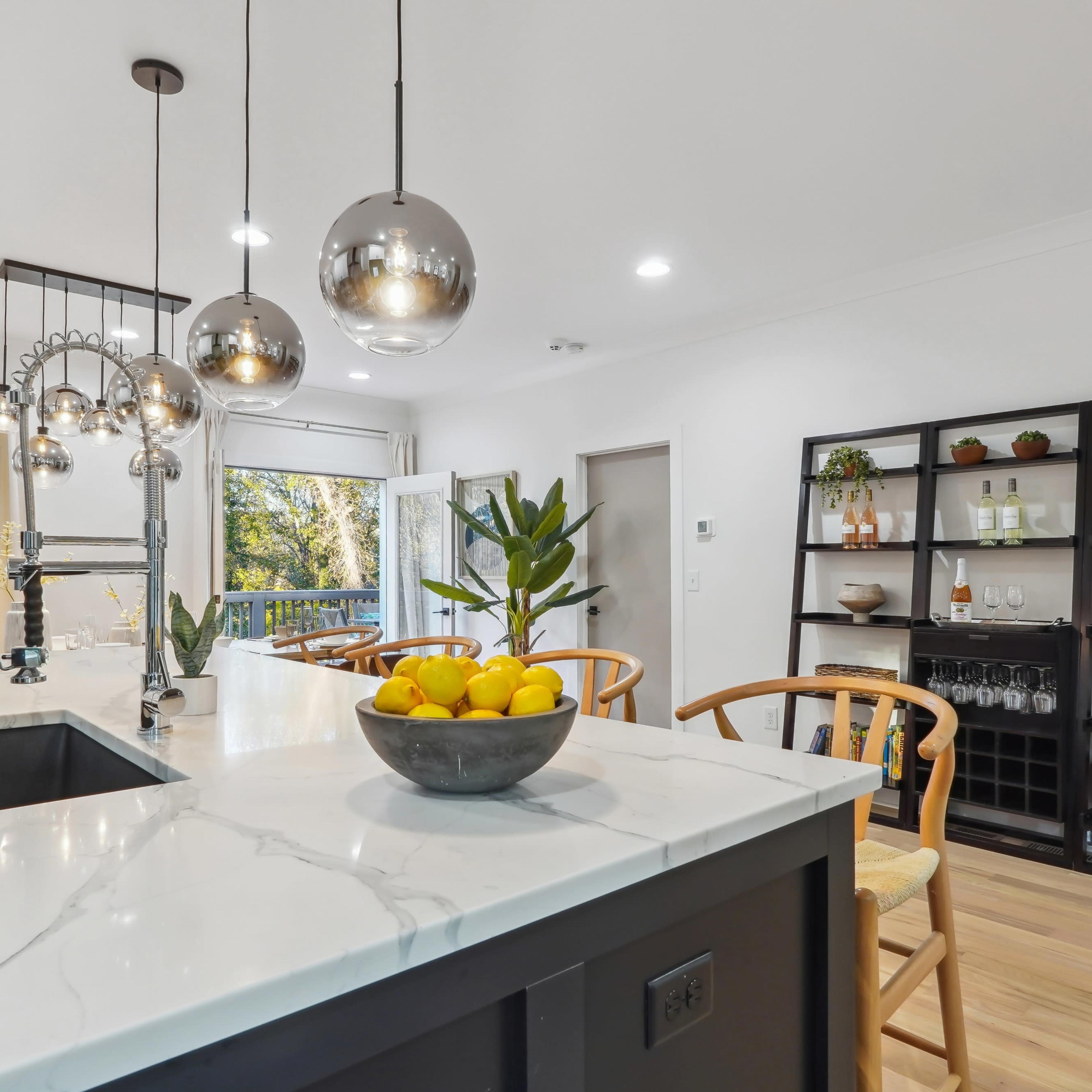 A modern kitchen with a large island featuring a bowl of lemons, pendant lights overhead, and a dining area in the background.