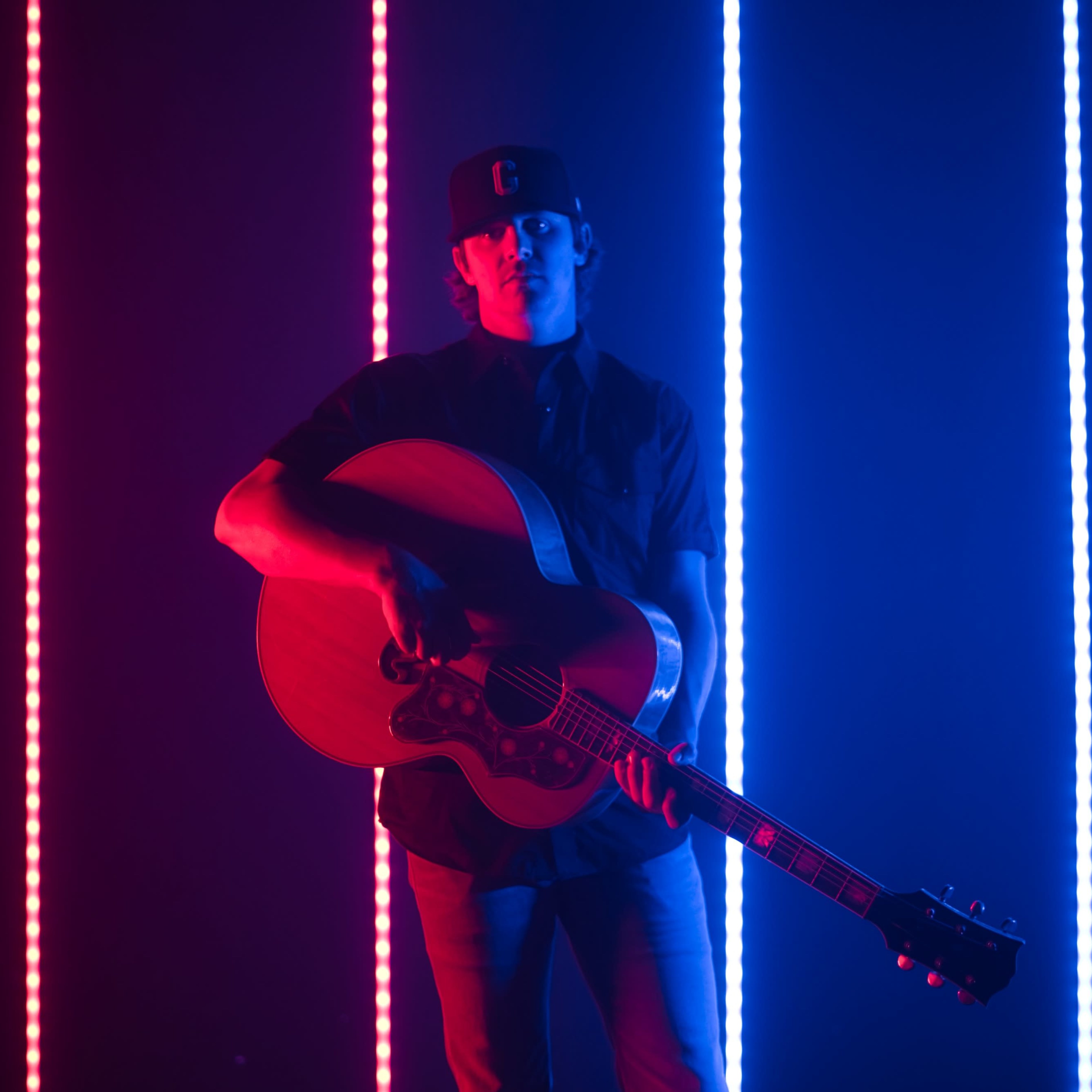 A musician stands holding an acoustic guitar in front of vertical light strips in red and blue.