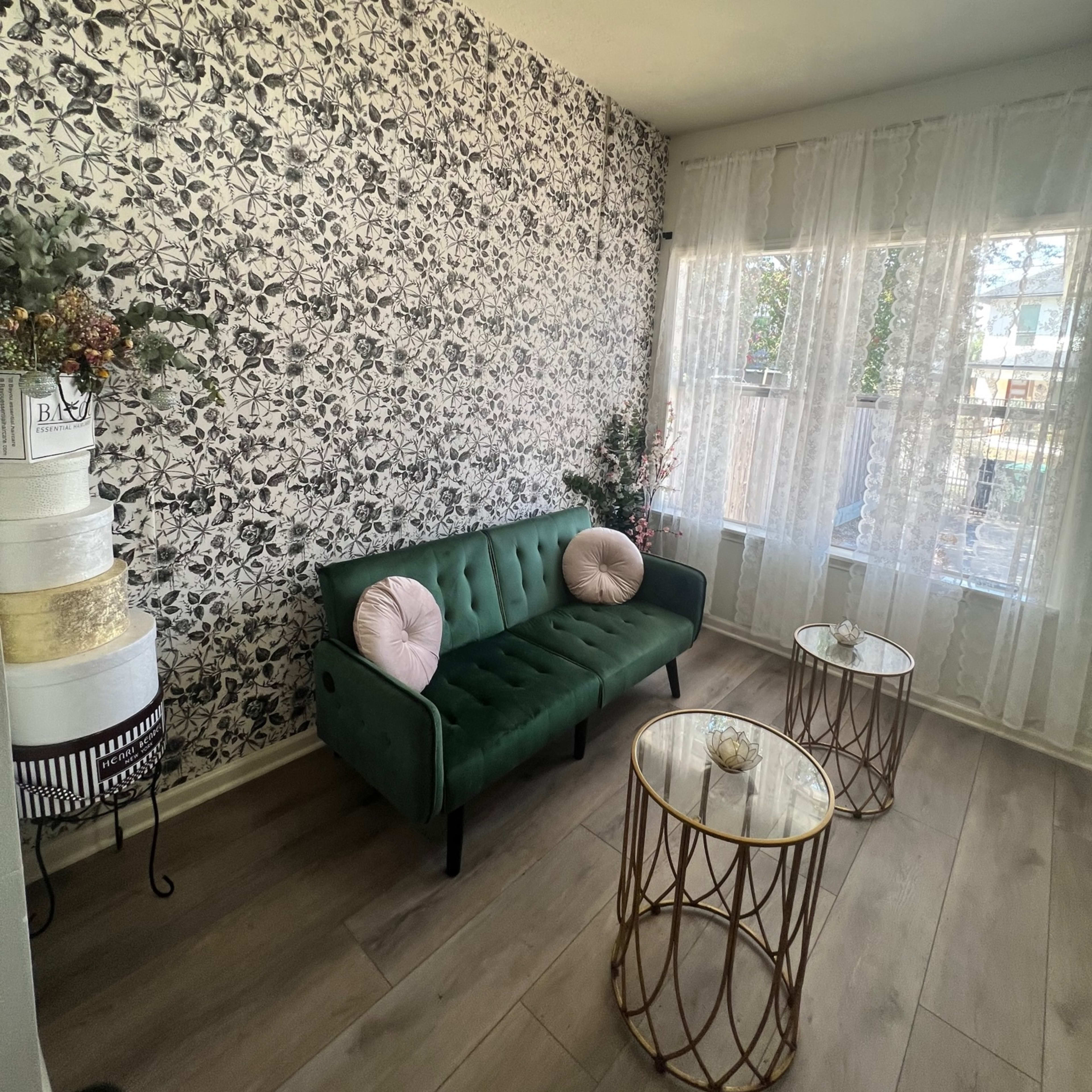 A stylish room with a green sofa, two round gold coffee tables, and a floral patterned wall, illuminated by a chandelier.