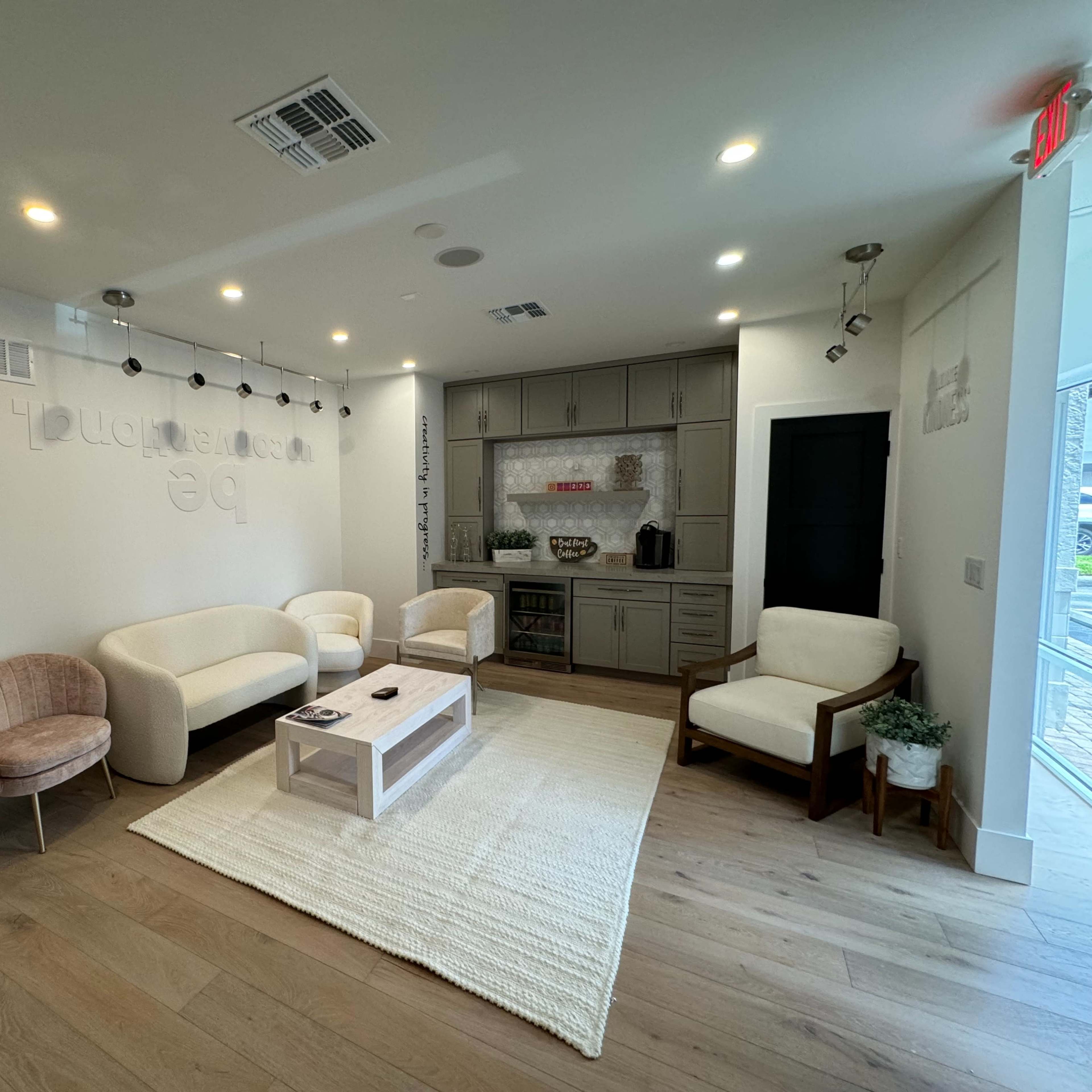 A modern waiting area featuring a light-colored sofa set, a coffee table, and a rug on a wooden floor, with a kitchenette visible in the background.