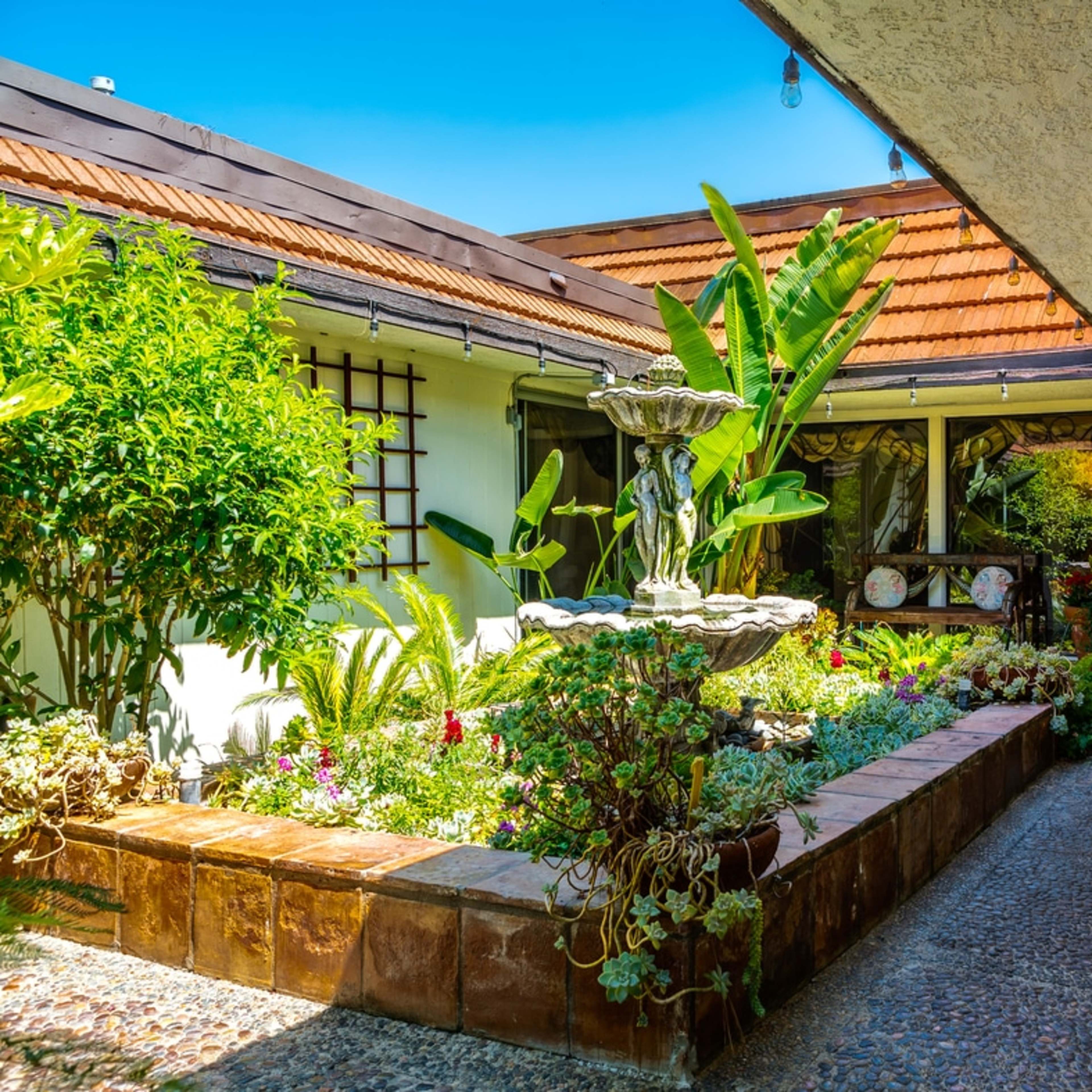 A pathway lined with greenery leads to a decorative fountain in a courtyard beside a house with a tiled roof.