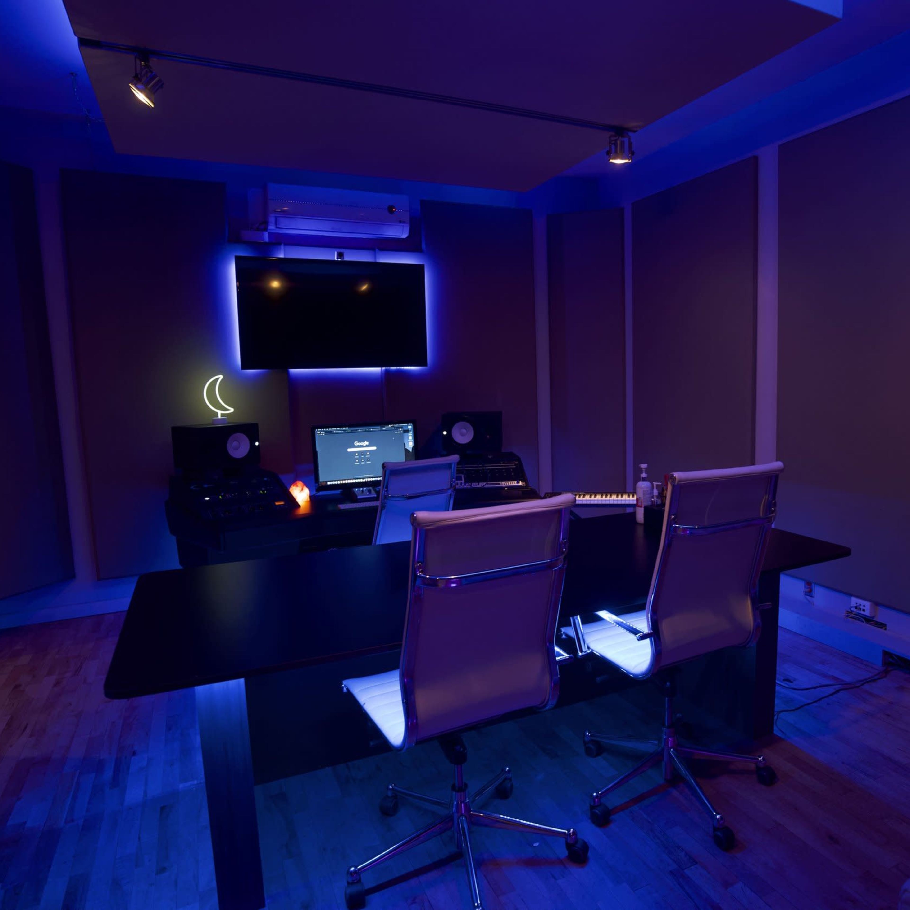 A music recording studio features a black table with two white chairs, surrounded by soundproof panels and illuminated by blue lighting.