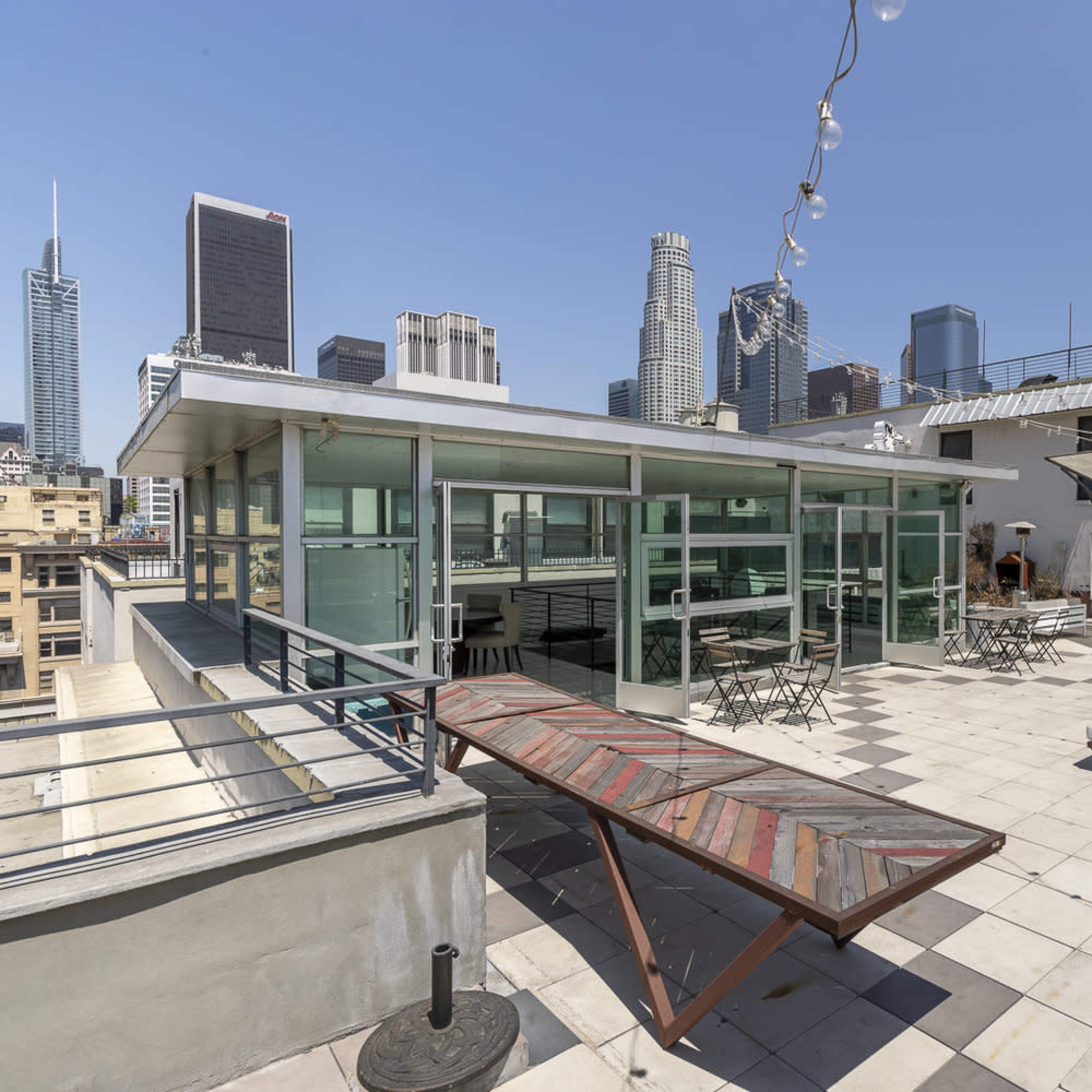 The rooftop features a spacious area with glass walls, tables, and lounge chairs, overlooking the downtown skyline.