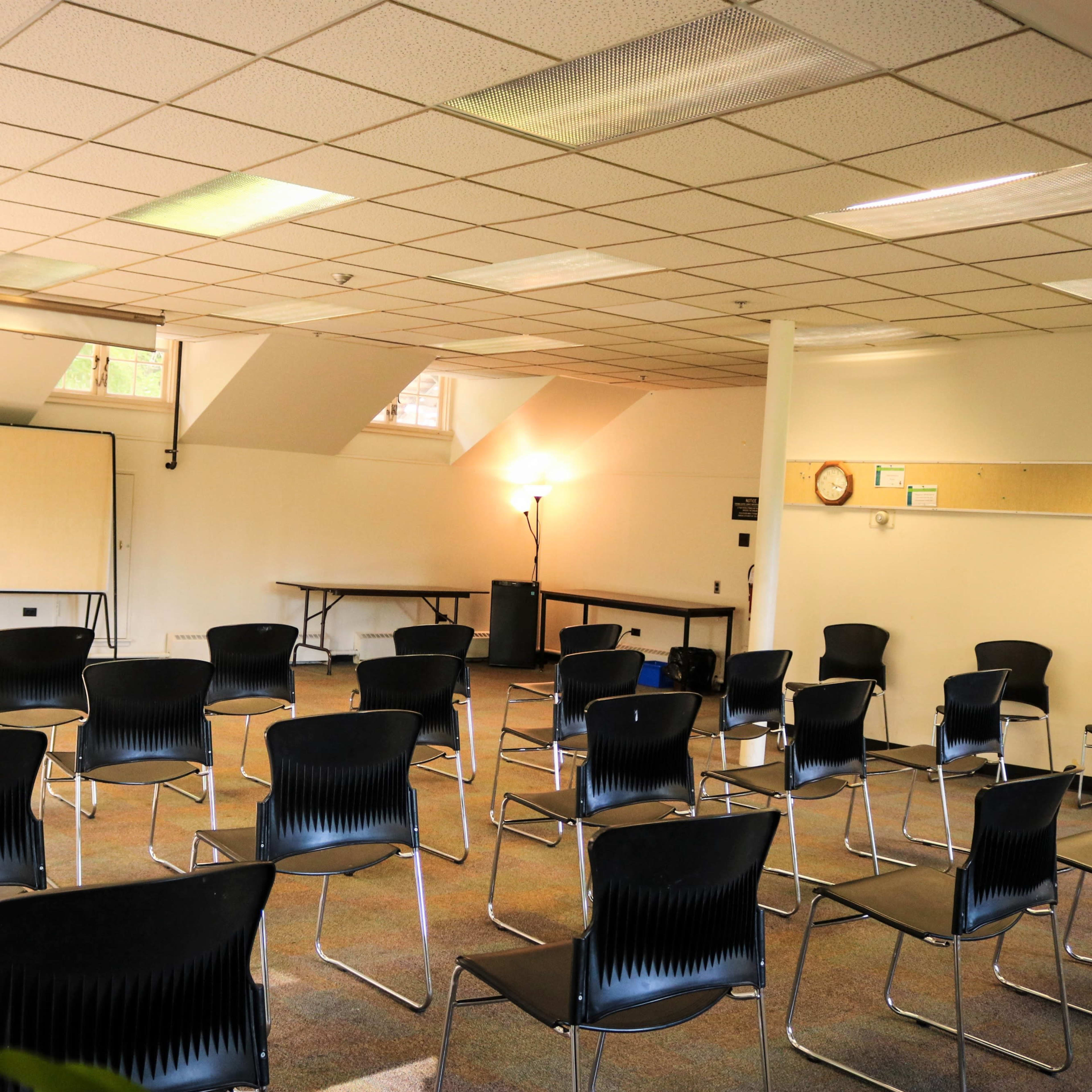 An empty meeting room with rows of black chairs arranged around a small table, a whiteboard on one side, and a large window allowing natural light.