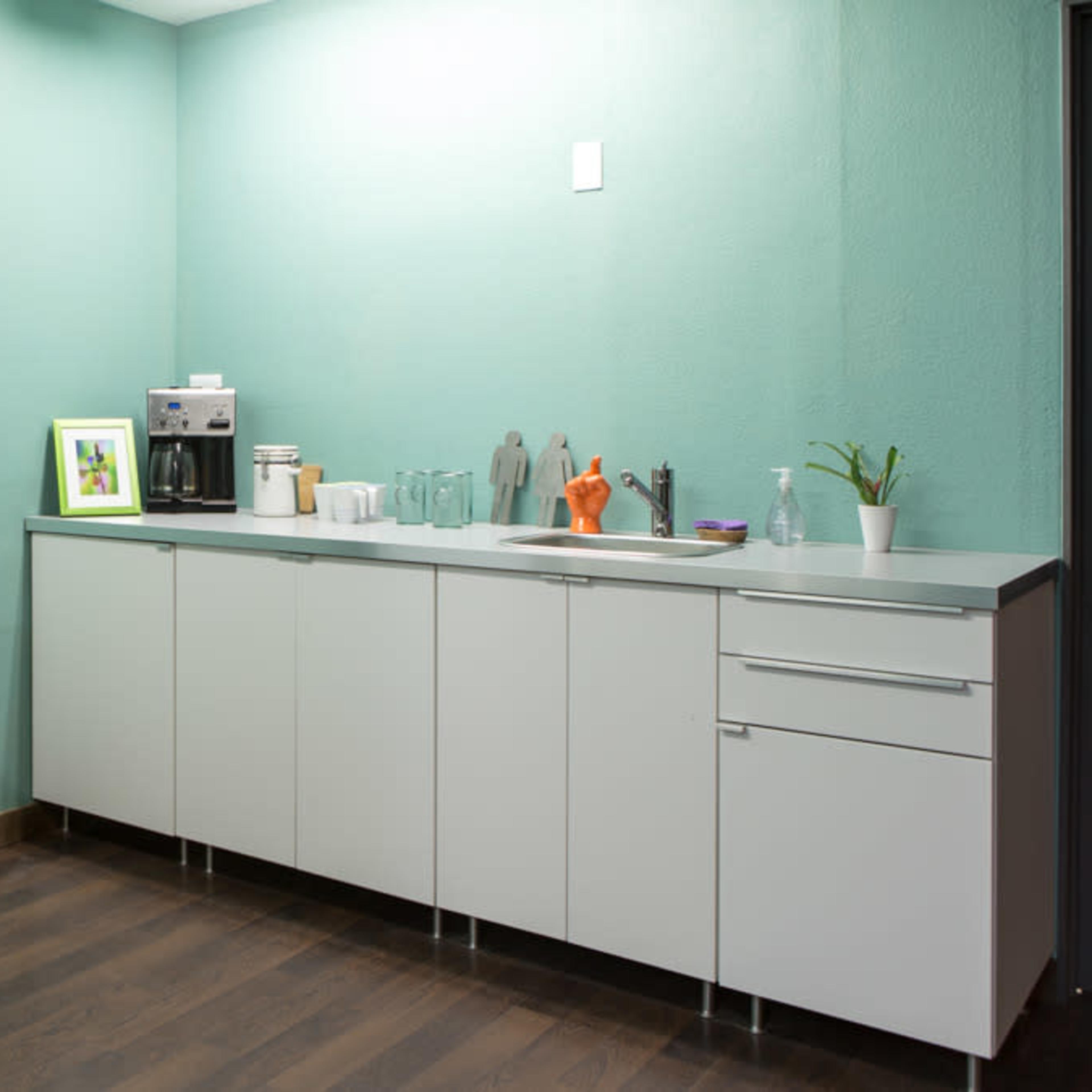 A modern kitchen area features mint green walls, a white cabinet, a sink, and a coffee maker, with a snack display on a stand.