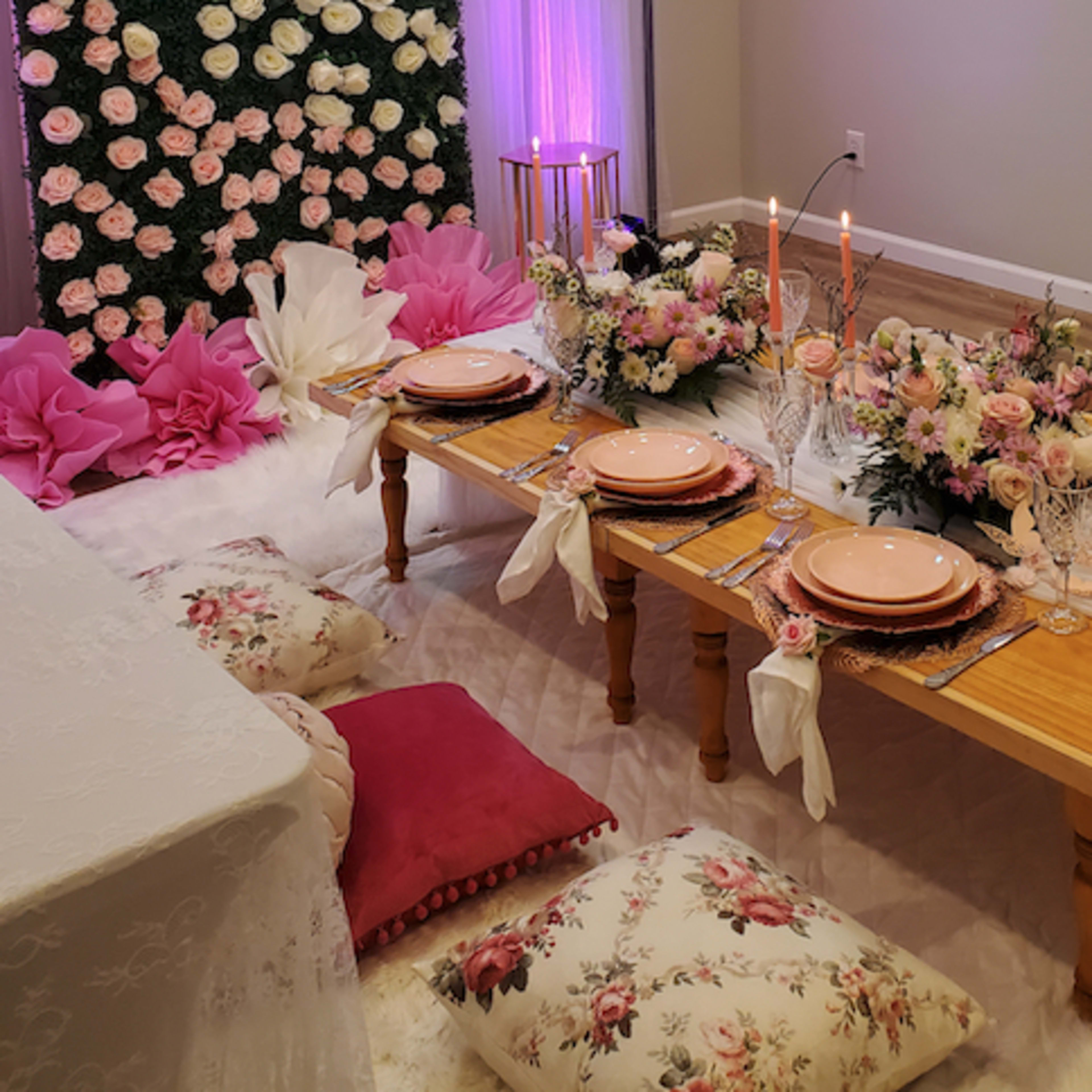 A decorated dining area with a low wooden table set for a meal, surrounded by floral arrangements and cushions on the floor.