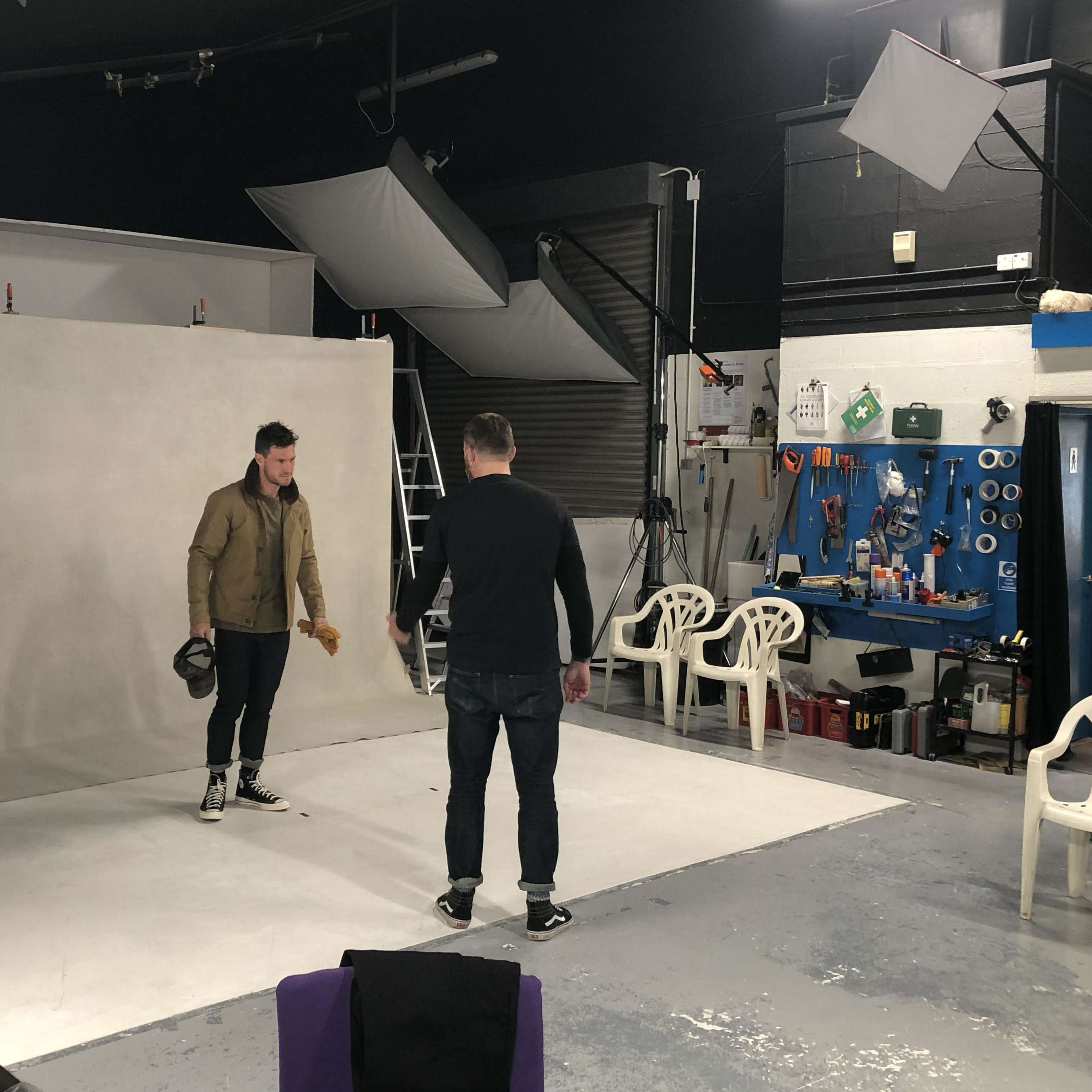 A photographer prepares to capture a subject dressed in a jacket and helmet in a well-equipped studio with a neutral backdrop.