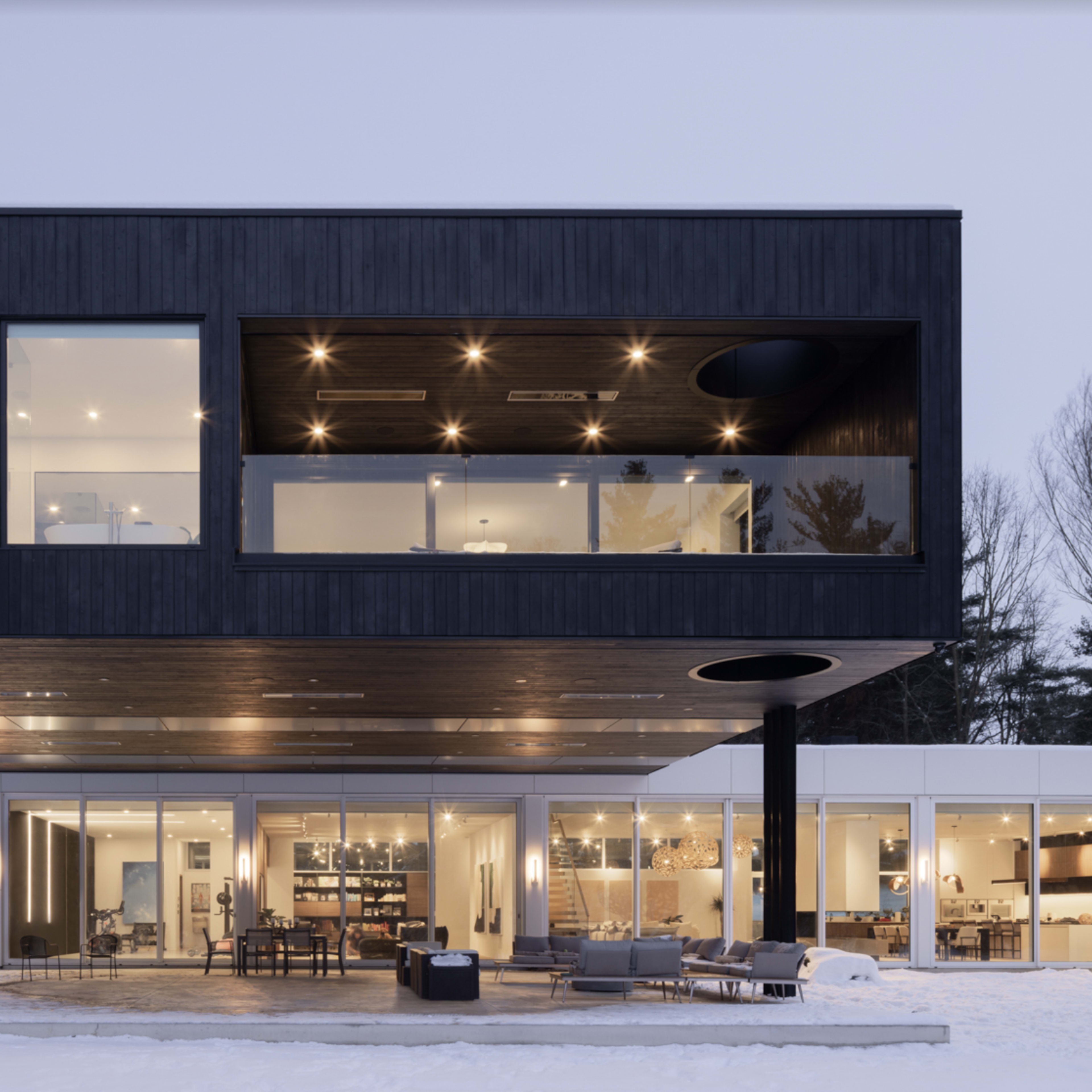 A modern two-story house with a dark wooden exterior and large windows overlooks a snowy landscape.