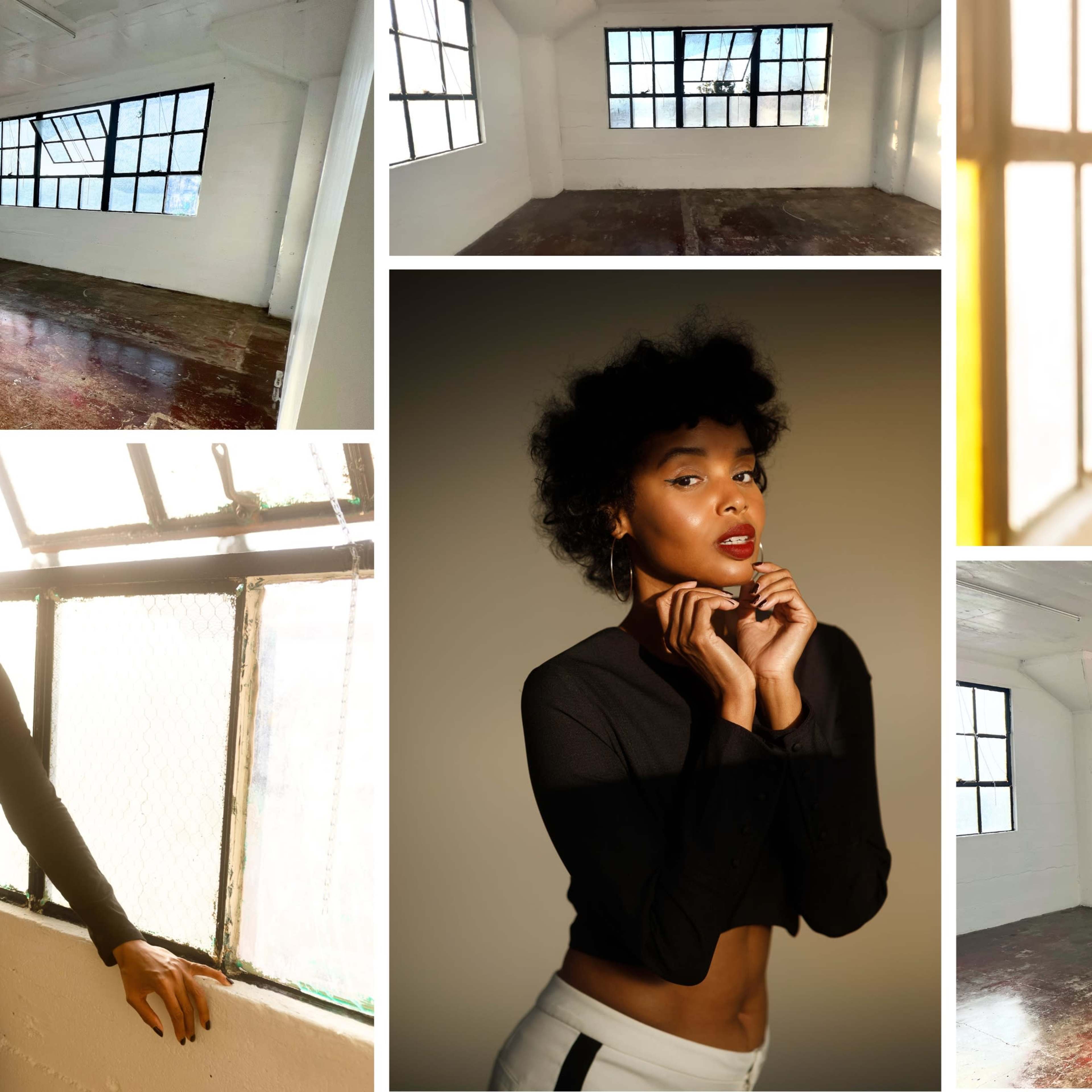 A model with curly hair posing in a brightly lit, empty studio room with large windows.