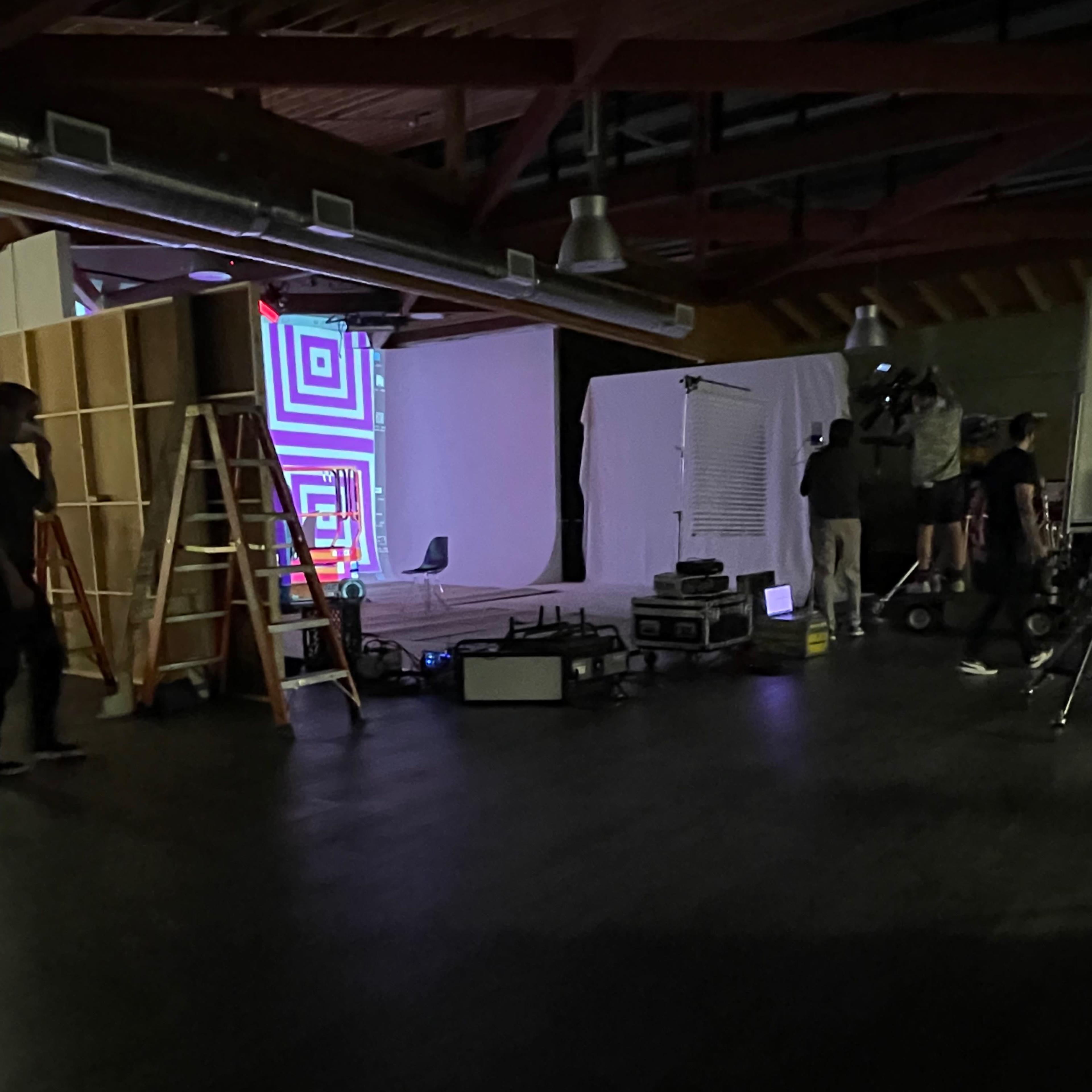A film or photo studio setup with equipment, including ladders and lighting, and a projected geometric pattern on a screen.