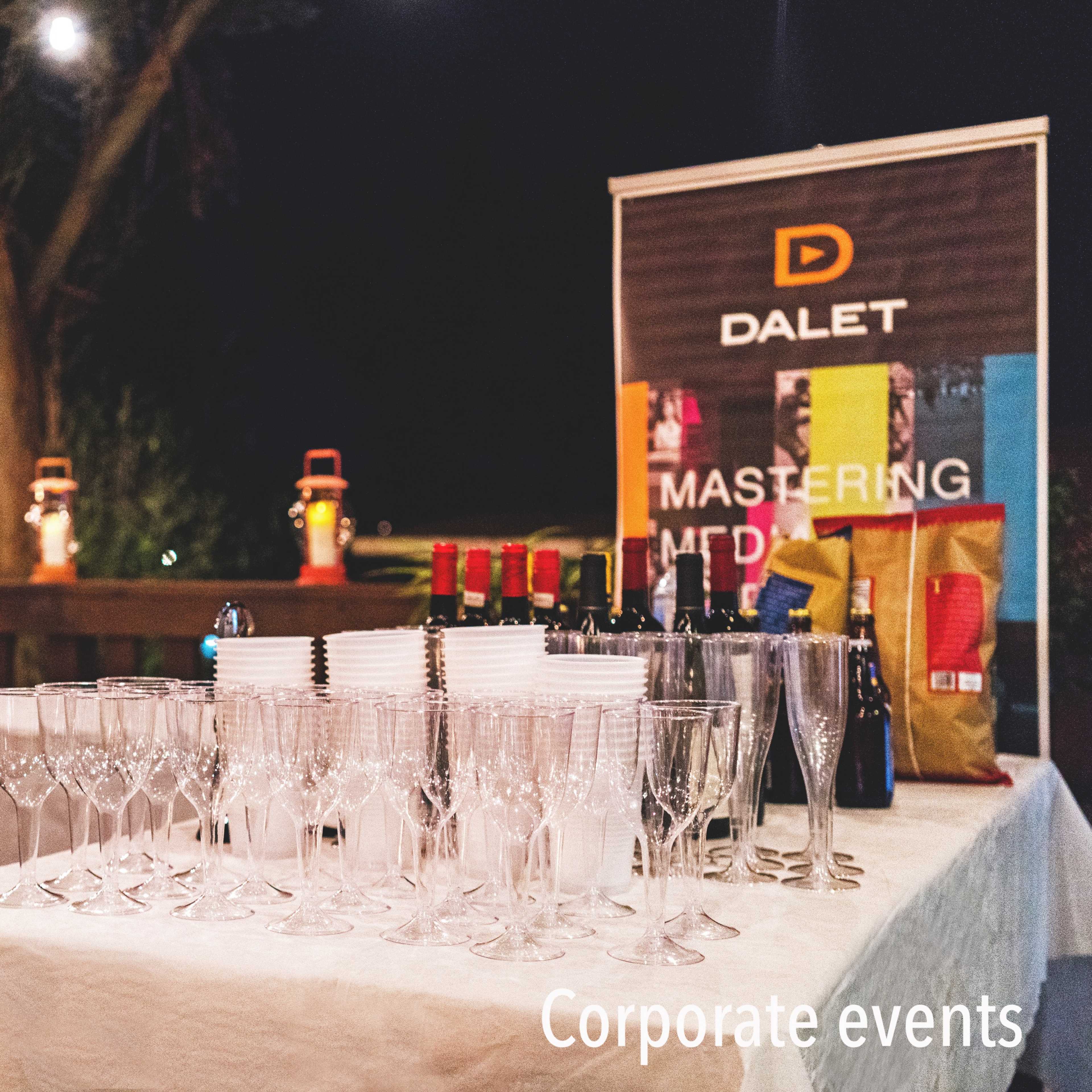 A table is set up outdoors at night with wine glasses, bottles, and a promotional banner for Dalet, alongside a few lit lanterns and string lights.