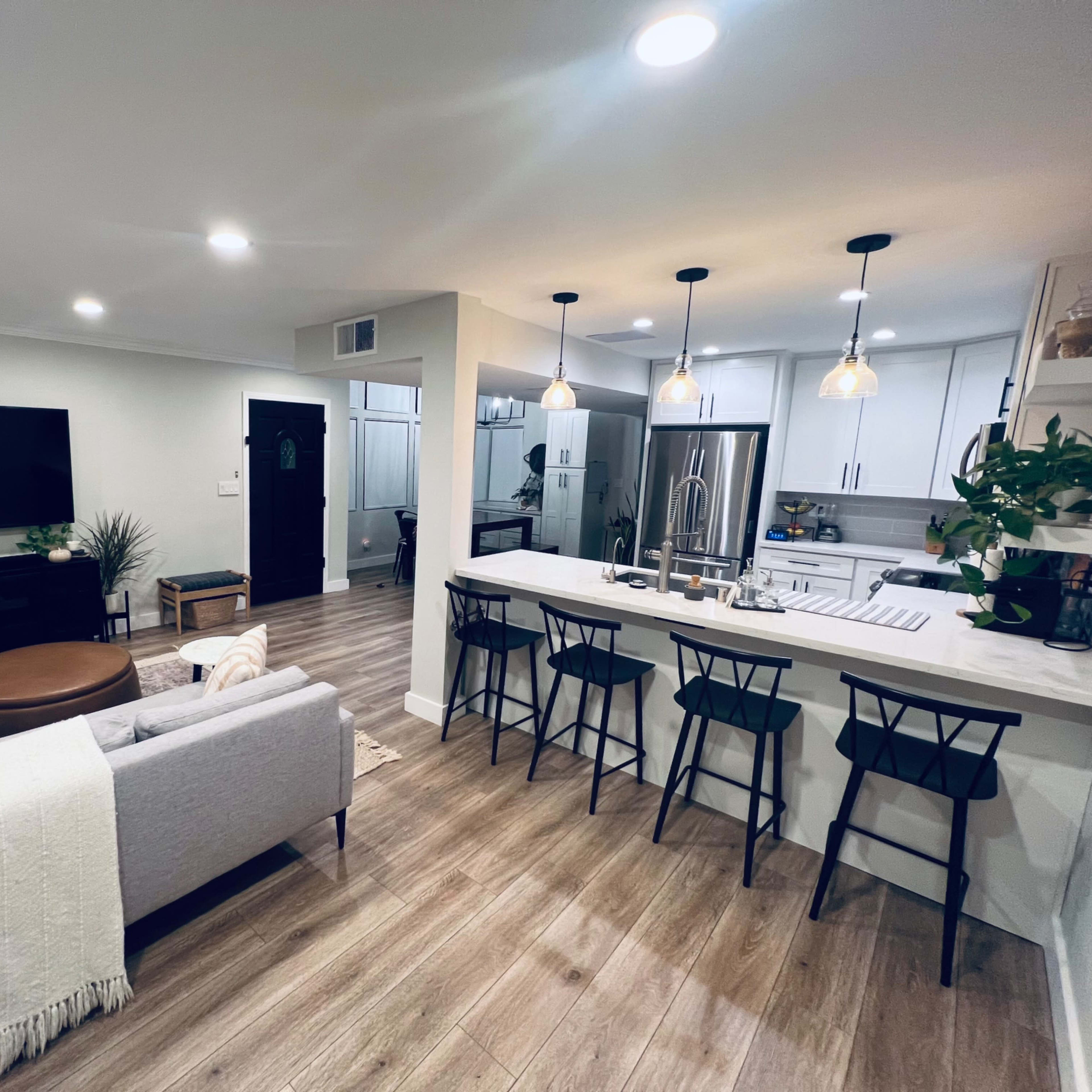 A modern living room and kitchen area with a gray sofa, a wooden coffee table, a large TV, and a kitchen counter with bar stools.