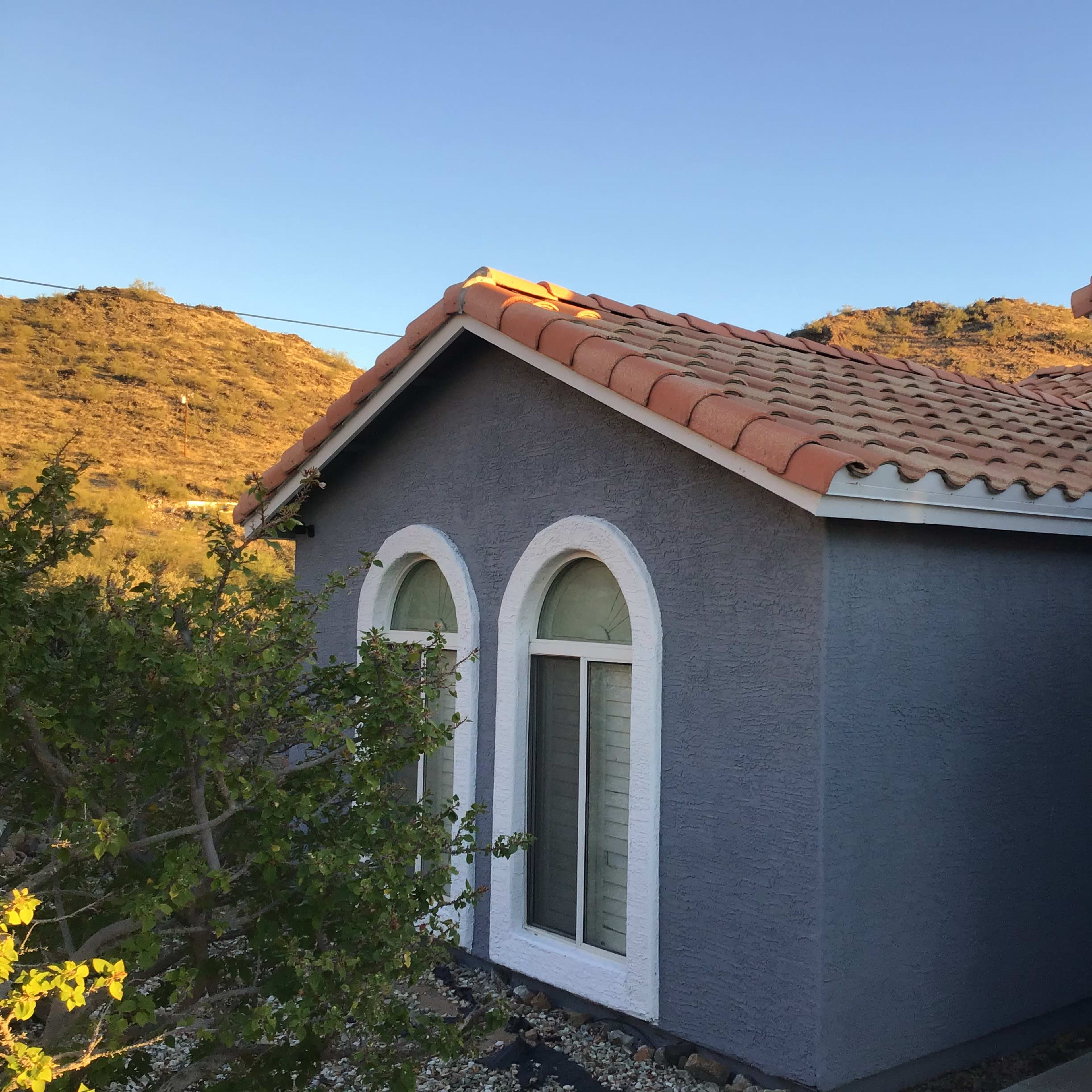 A single-story house with a gray exterior, arched windows, and a tiled roof, situated near a hillside.
