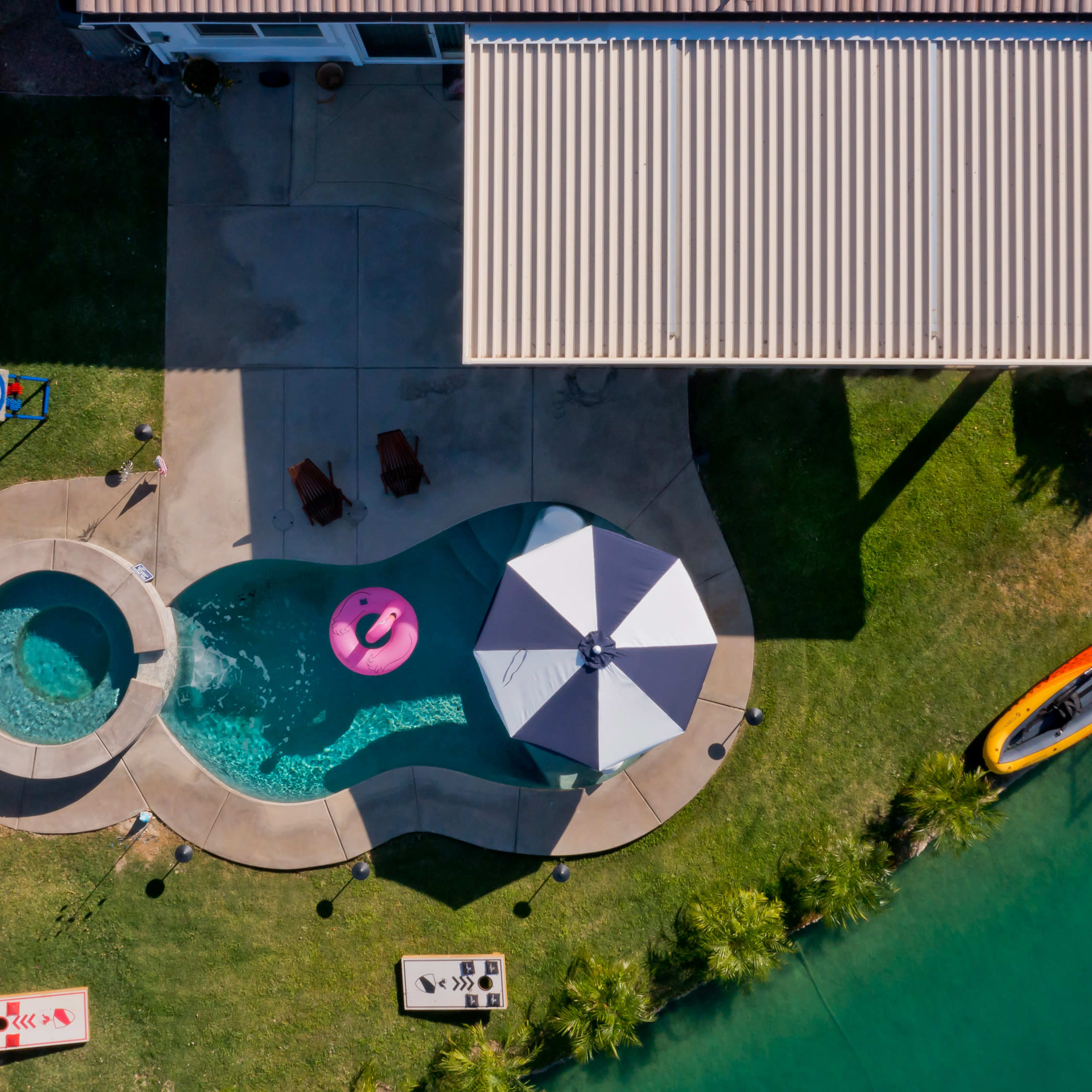 An aerial view of a backyard with a swimming pool, hot tub, patio umbrella, two lounge chairs, and a kayaks by a water body.