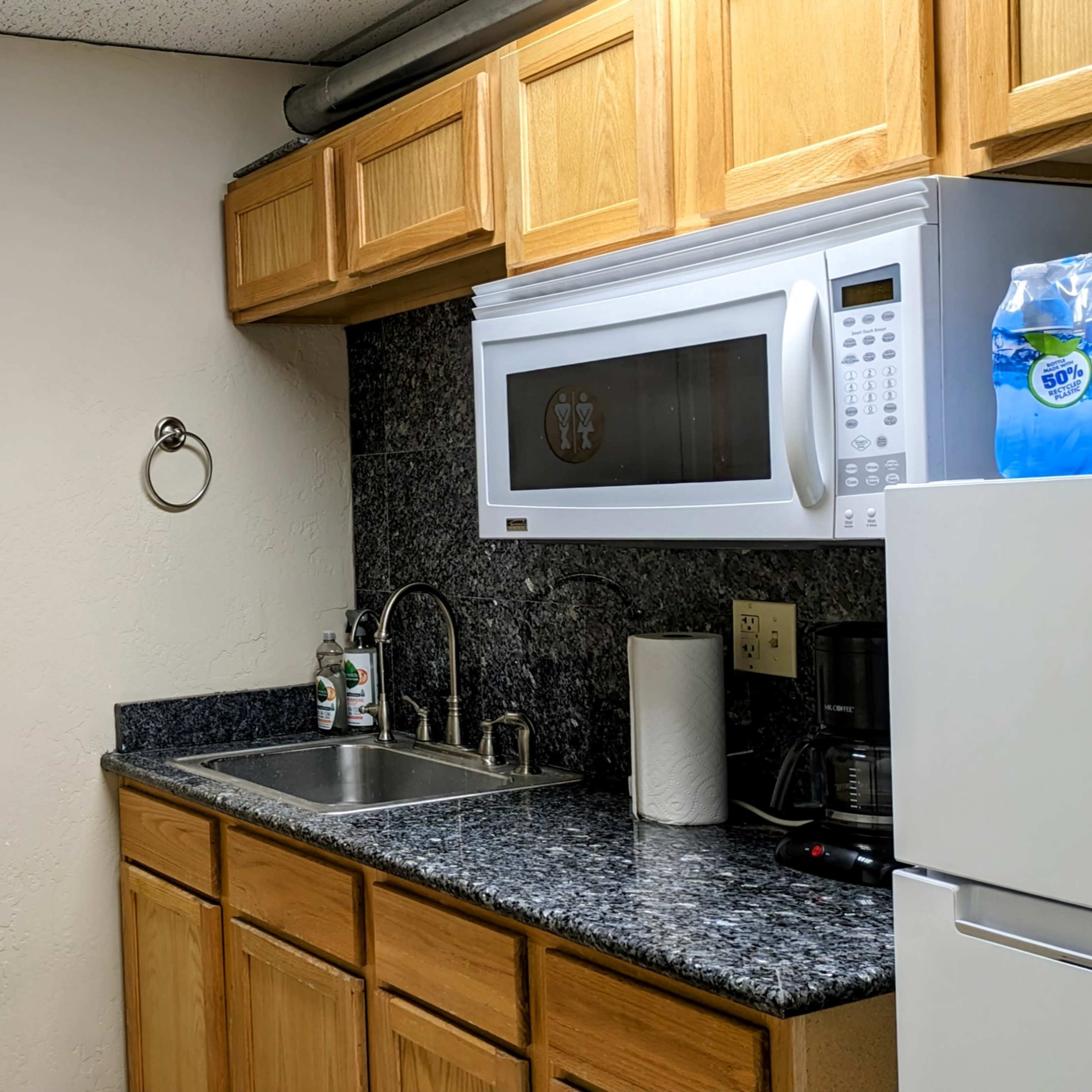 A small kitchen area with wooden cabinets, a sink, a microwave, and a closed door featuring a decorative emblem.