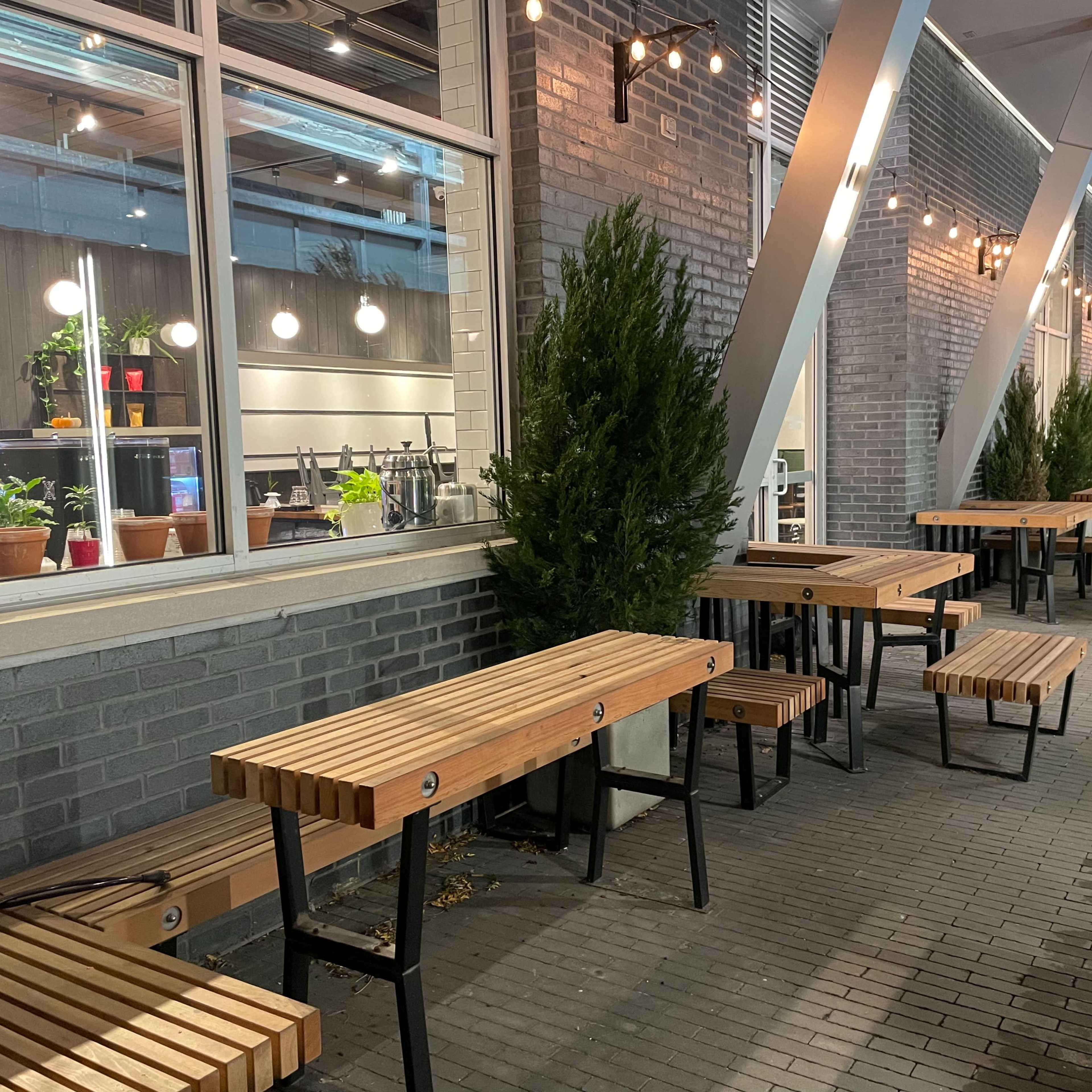 A row of wooden tables lined up along a brick wall, with potted plants and bright lights above.