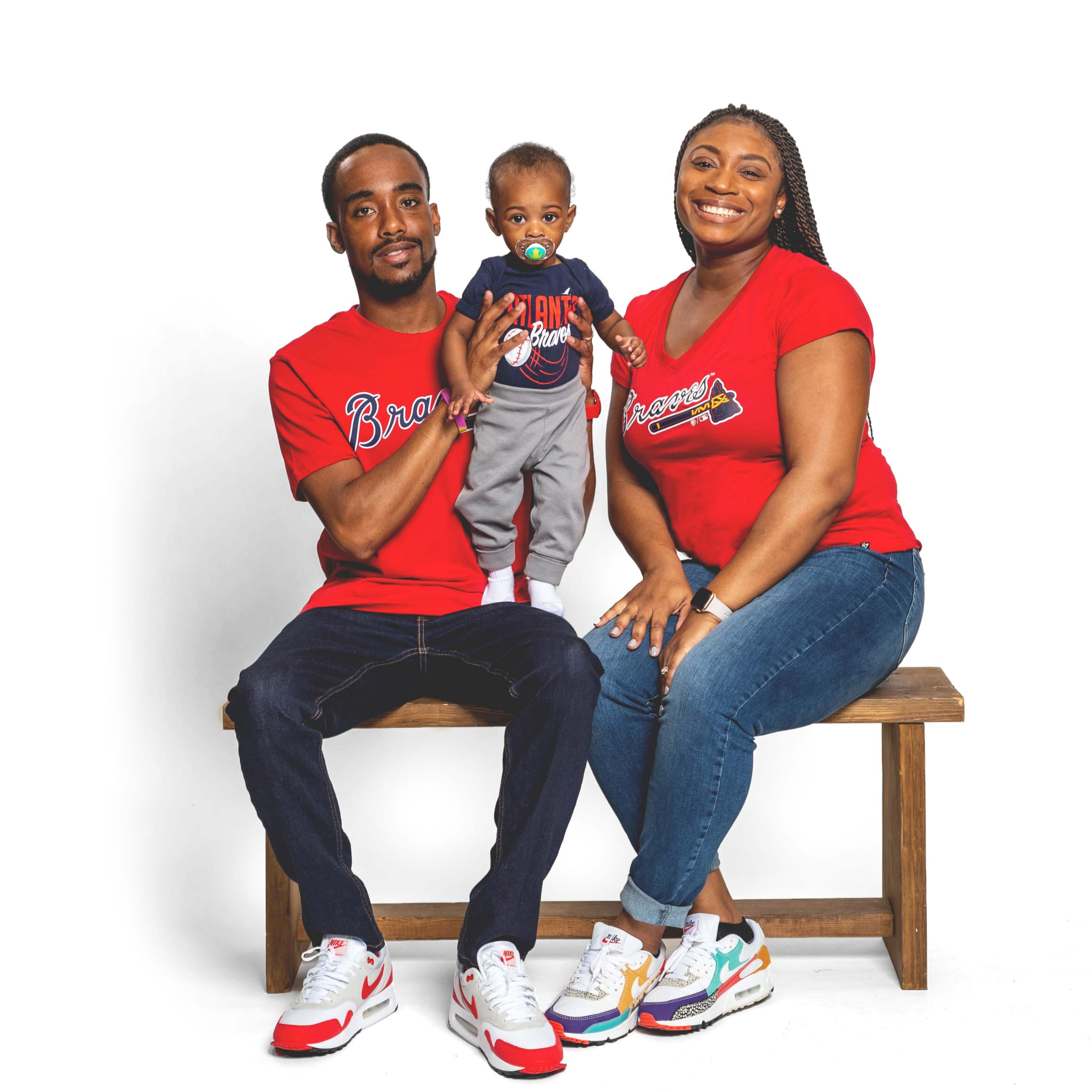 A man and a woman sit on a wooden bench, holding a baby dressed in gray, all wearing matching red shirts with a logo.