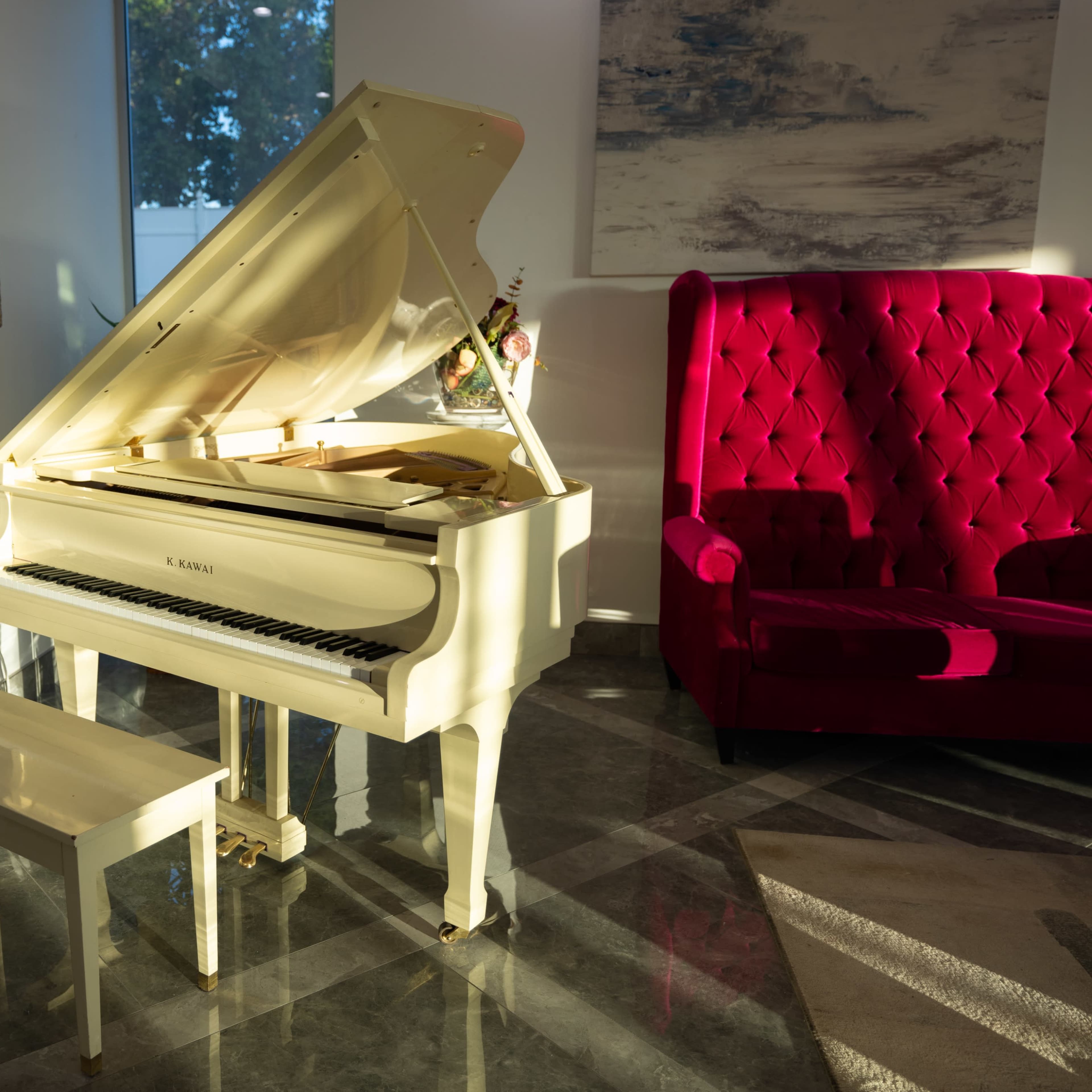 A white grand piano sits beside a red velvet armchair in a well-lit room with modern decor.