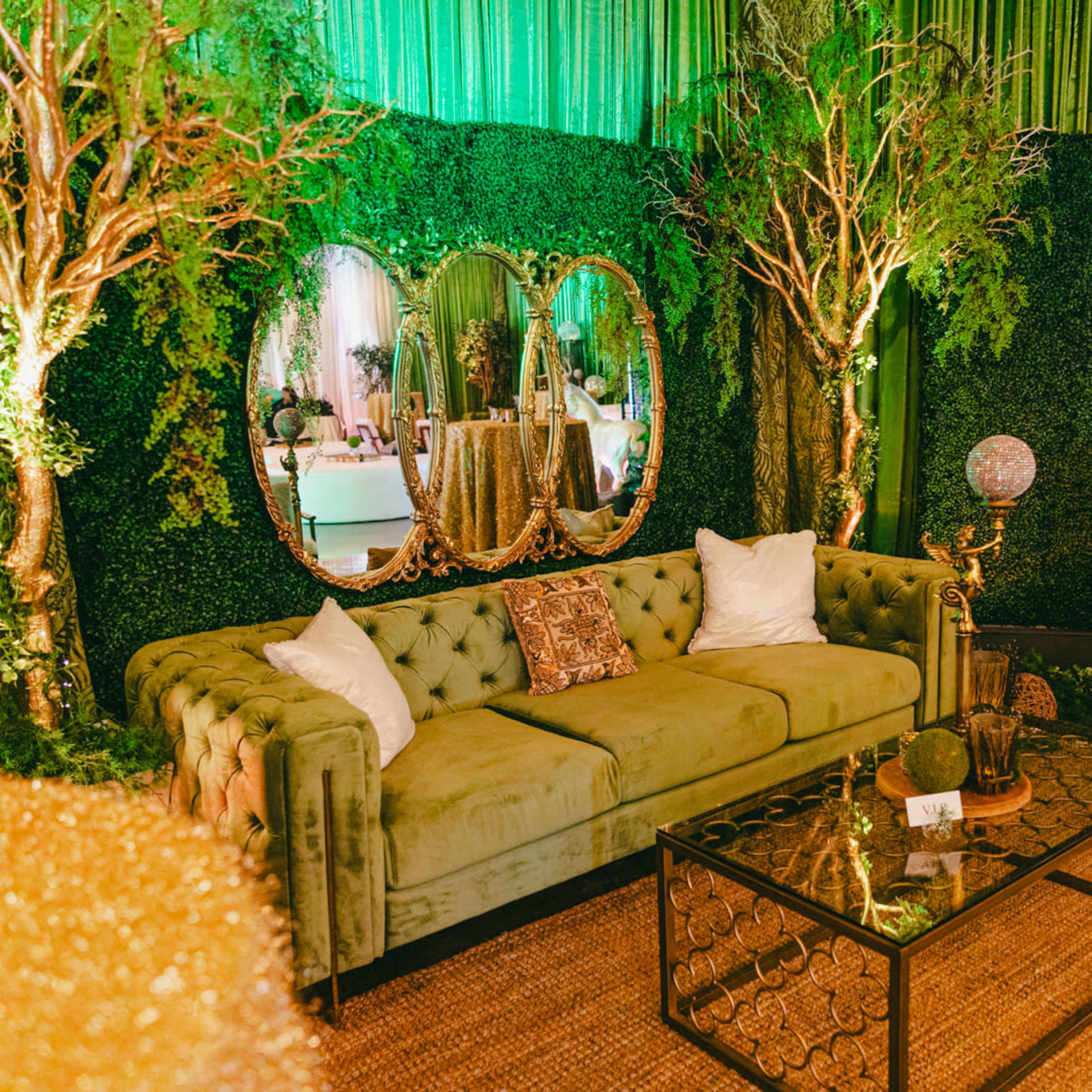 A decorated lounge area with a green velvet sofa, a glass coffee table, and lush greenery surrounding a mirror in an elegantly styled setting.