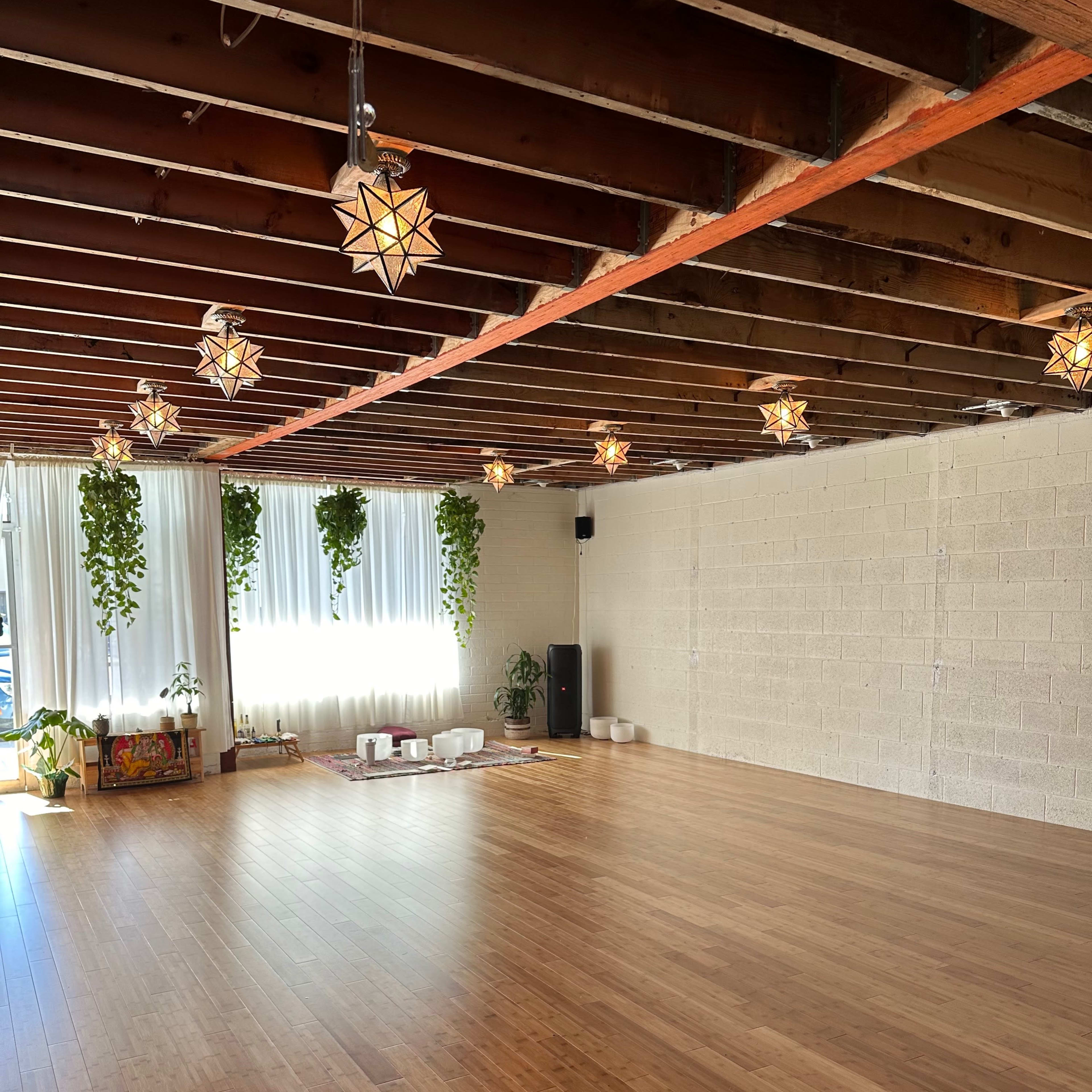 A spacious wooden yoga studio with overhead lights and plants along the walls.