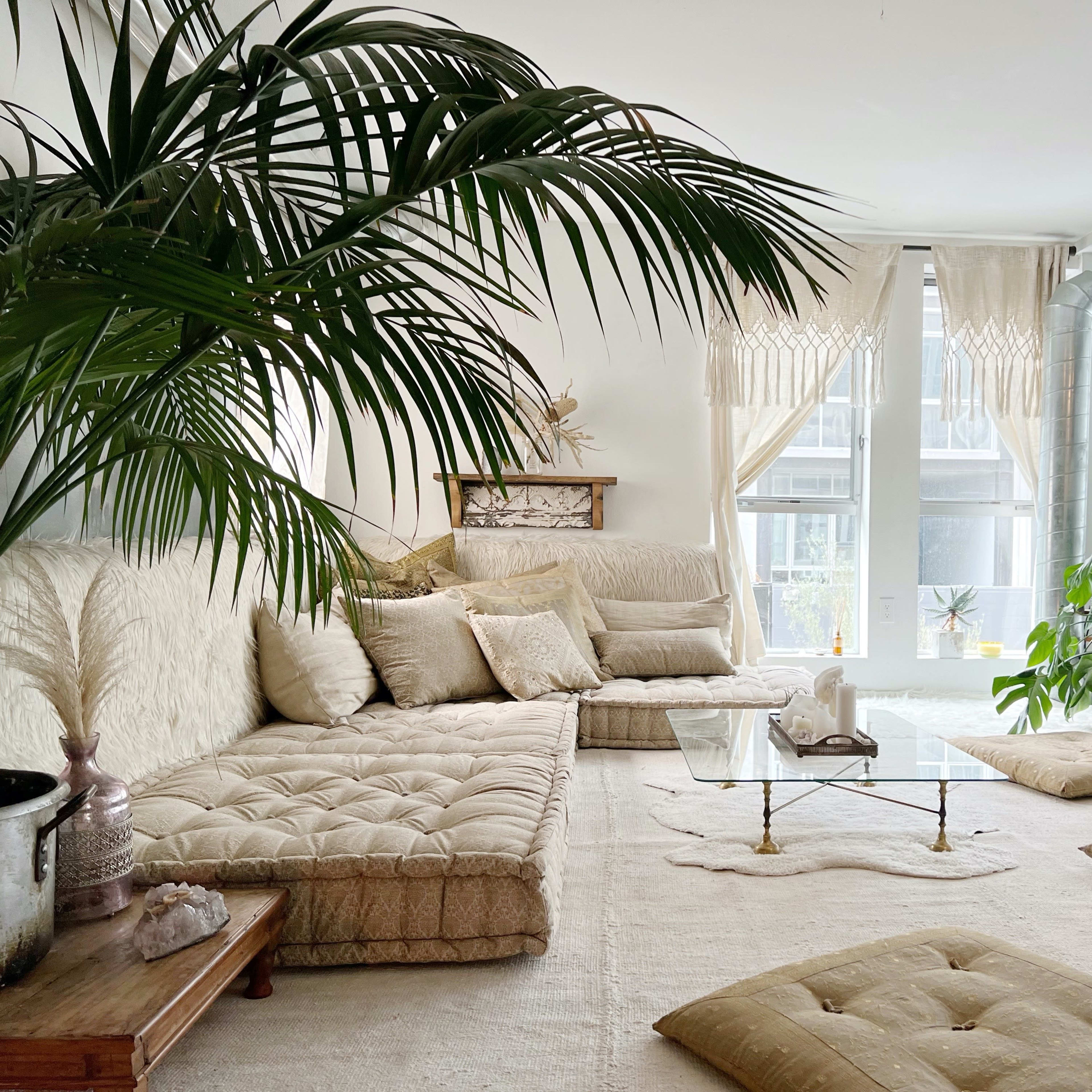 The room features a spacious, light-filled living area with a large, plush sectional sofa, several floor cushions, a glass coffee table, and an abundance of indoor plants.