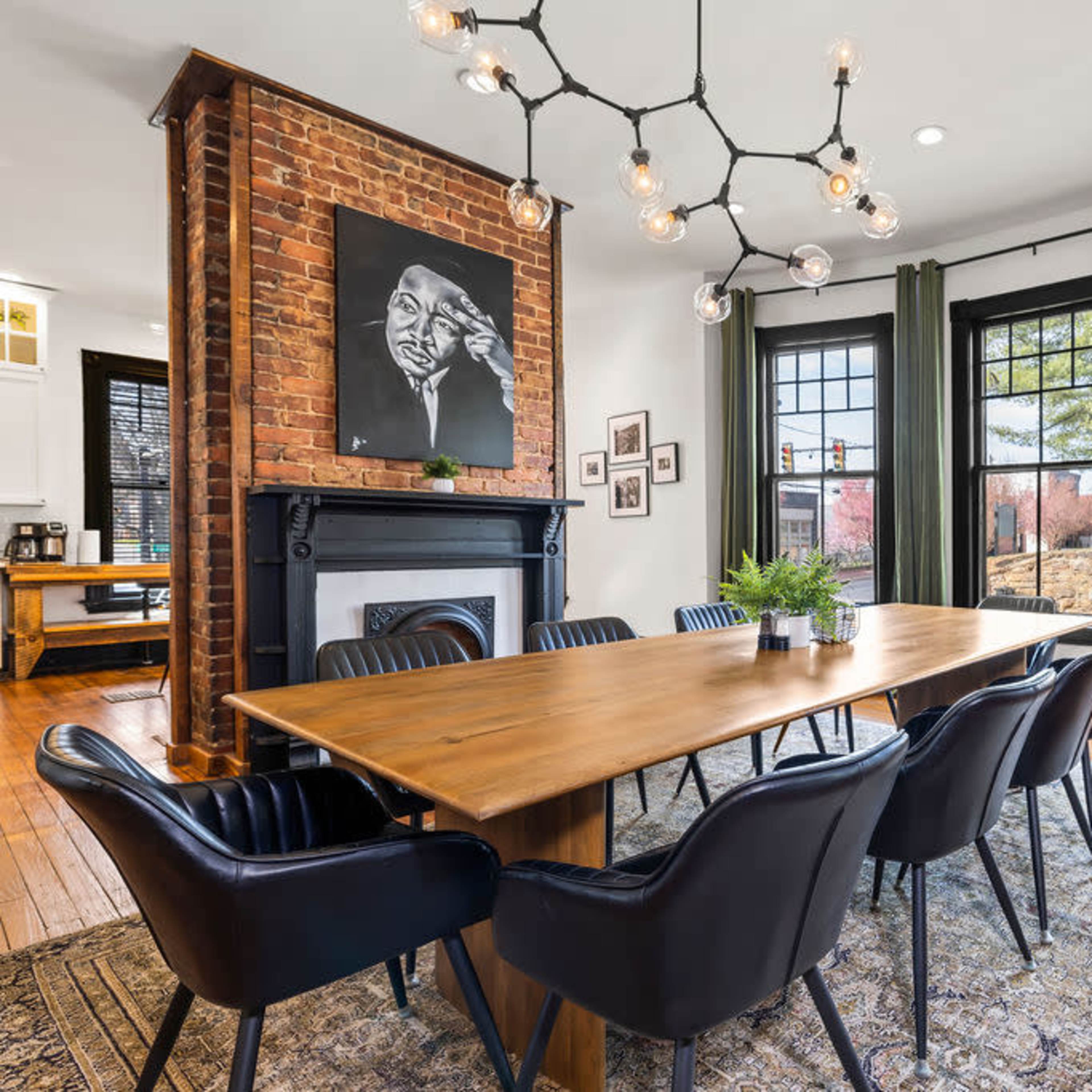 A dining room featuring a long wooden table surrounded by black chairs, with a large brick fireplace, artwork on the wall, and large windows dressed in green curtains.