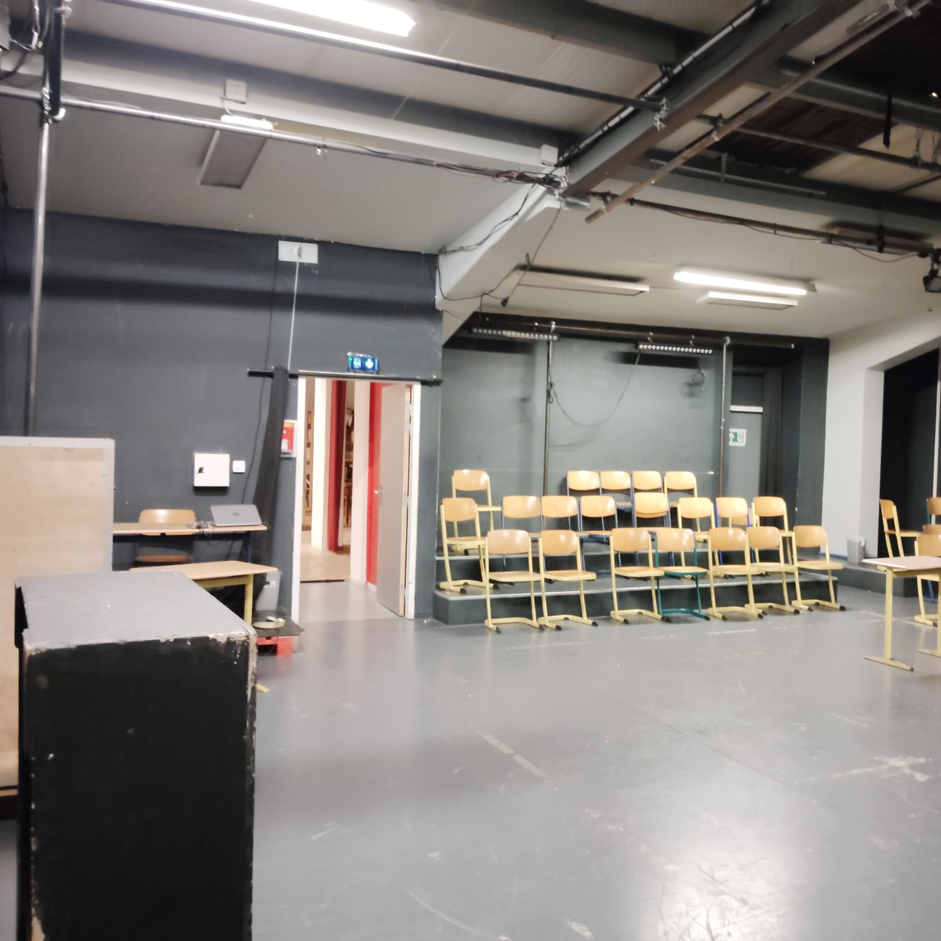 An empty rehearsal space with rows of wooden chairs facing a stage area and a doorway leading to a hallway.